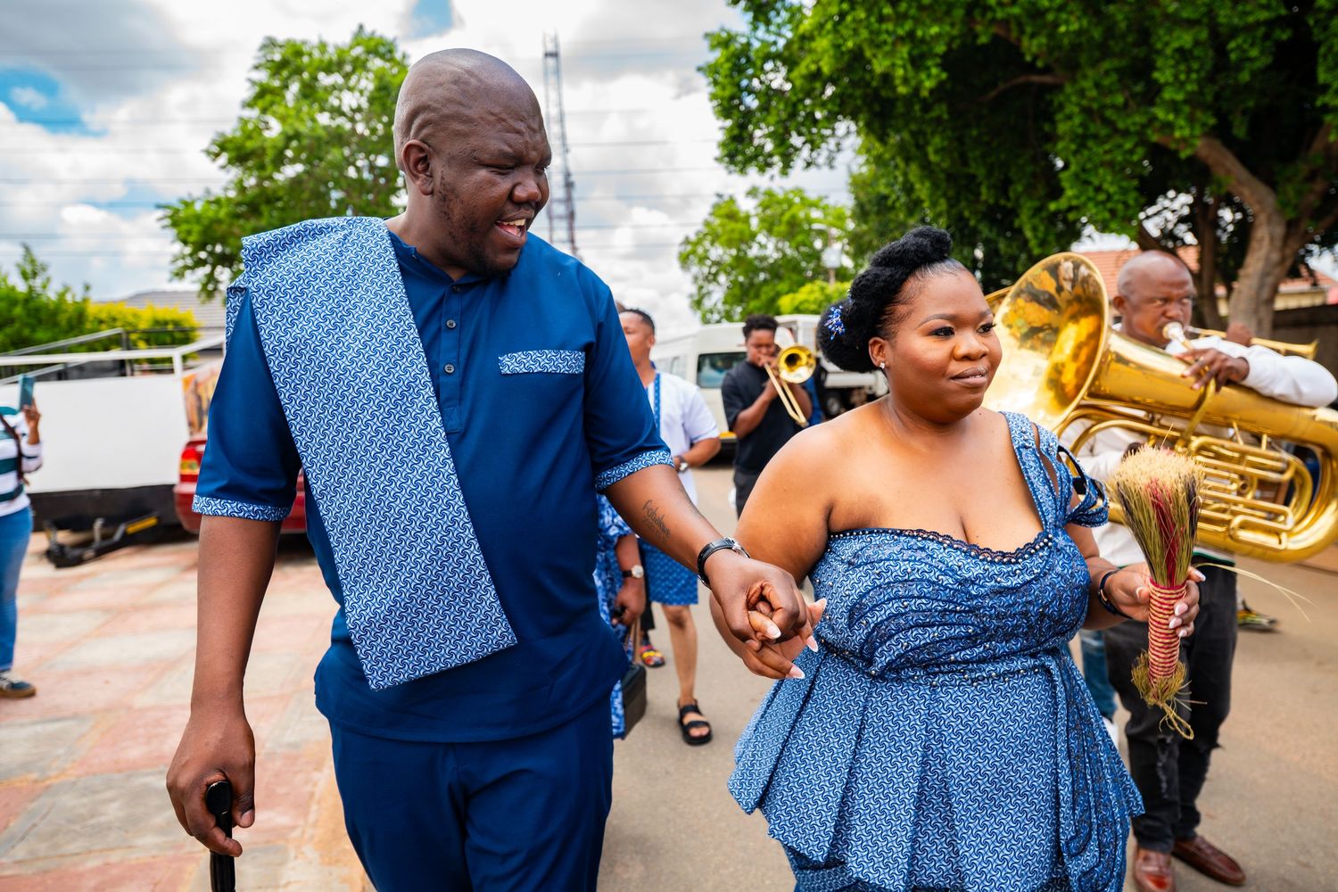 Couple in matching blue traditional African attire walking hand in hand at an outdoor celebration with brass band.