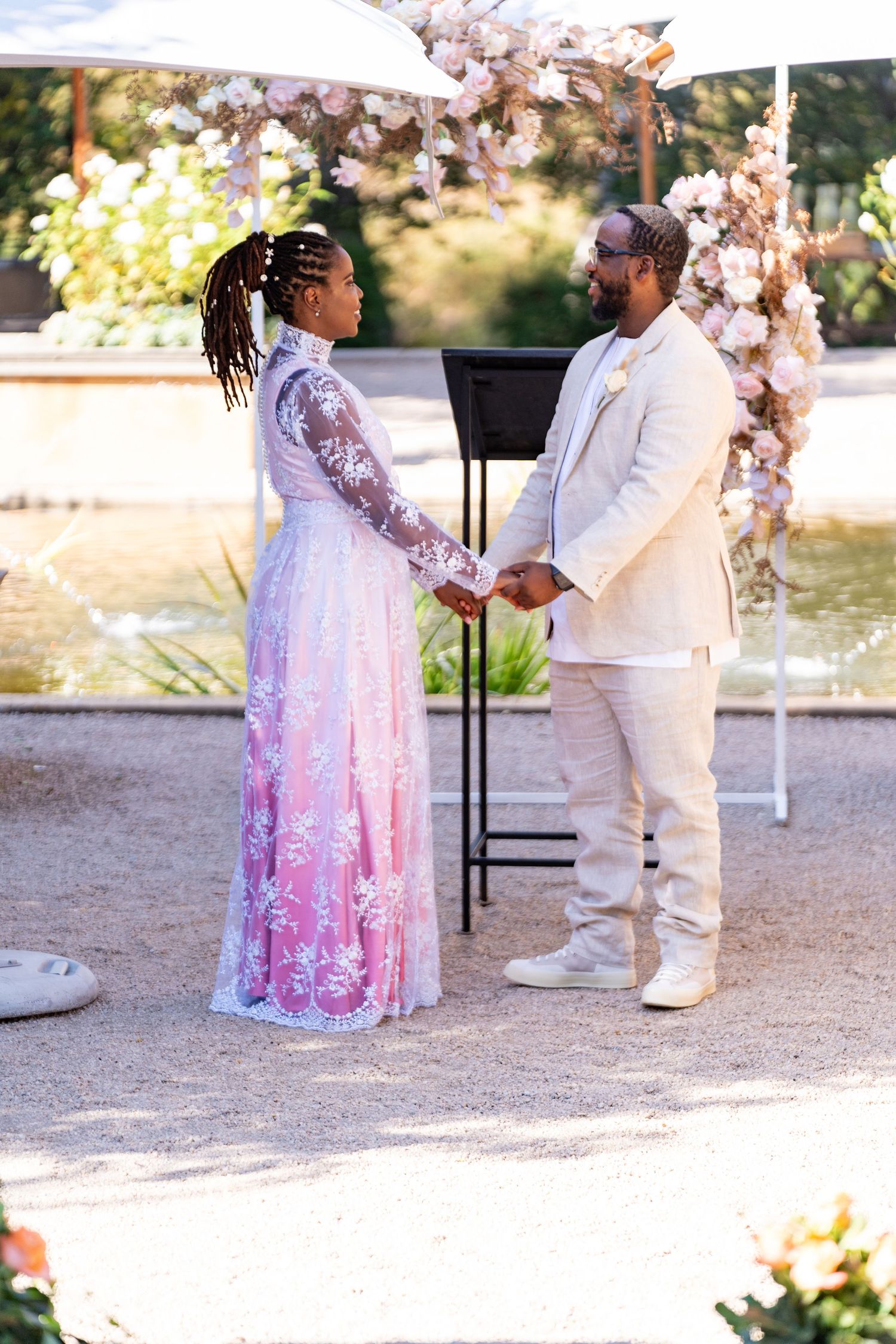 Couple exchanging vows under a floral arch by a pond, bride in a pink lace wedding gown.