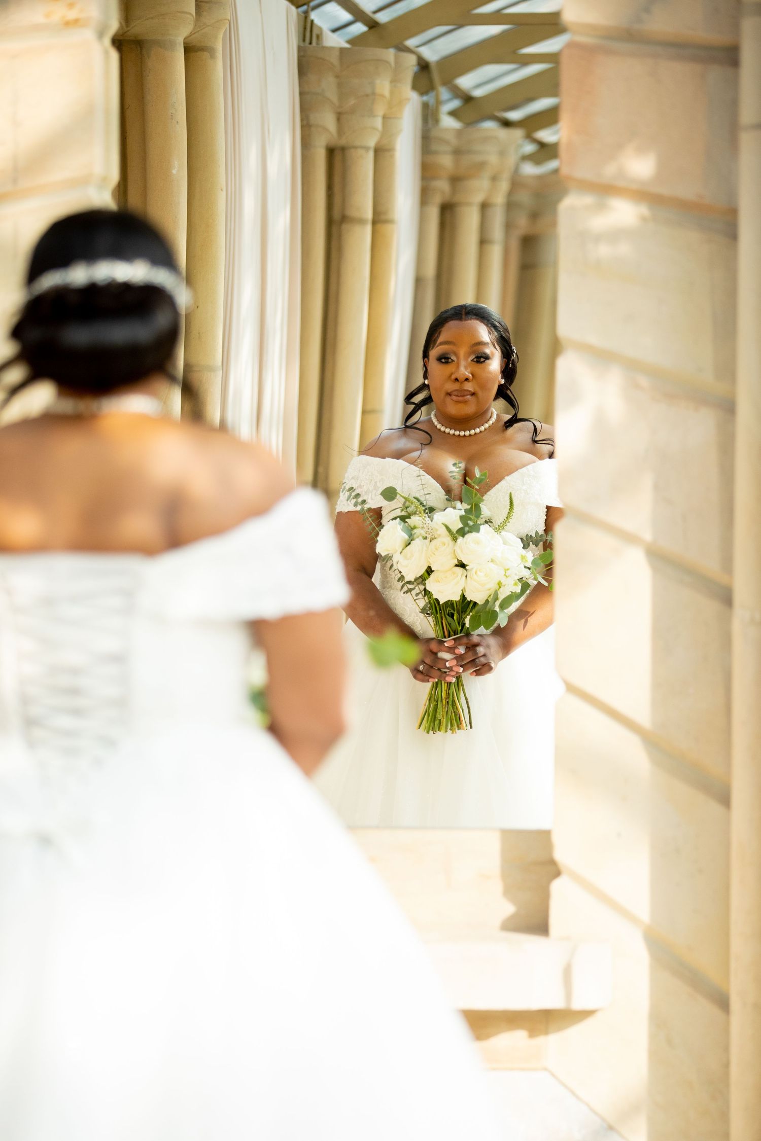 Bridesmaid in white dress holding bouquet gazing at bride in off-shoulder gown between columns.