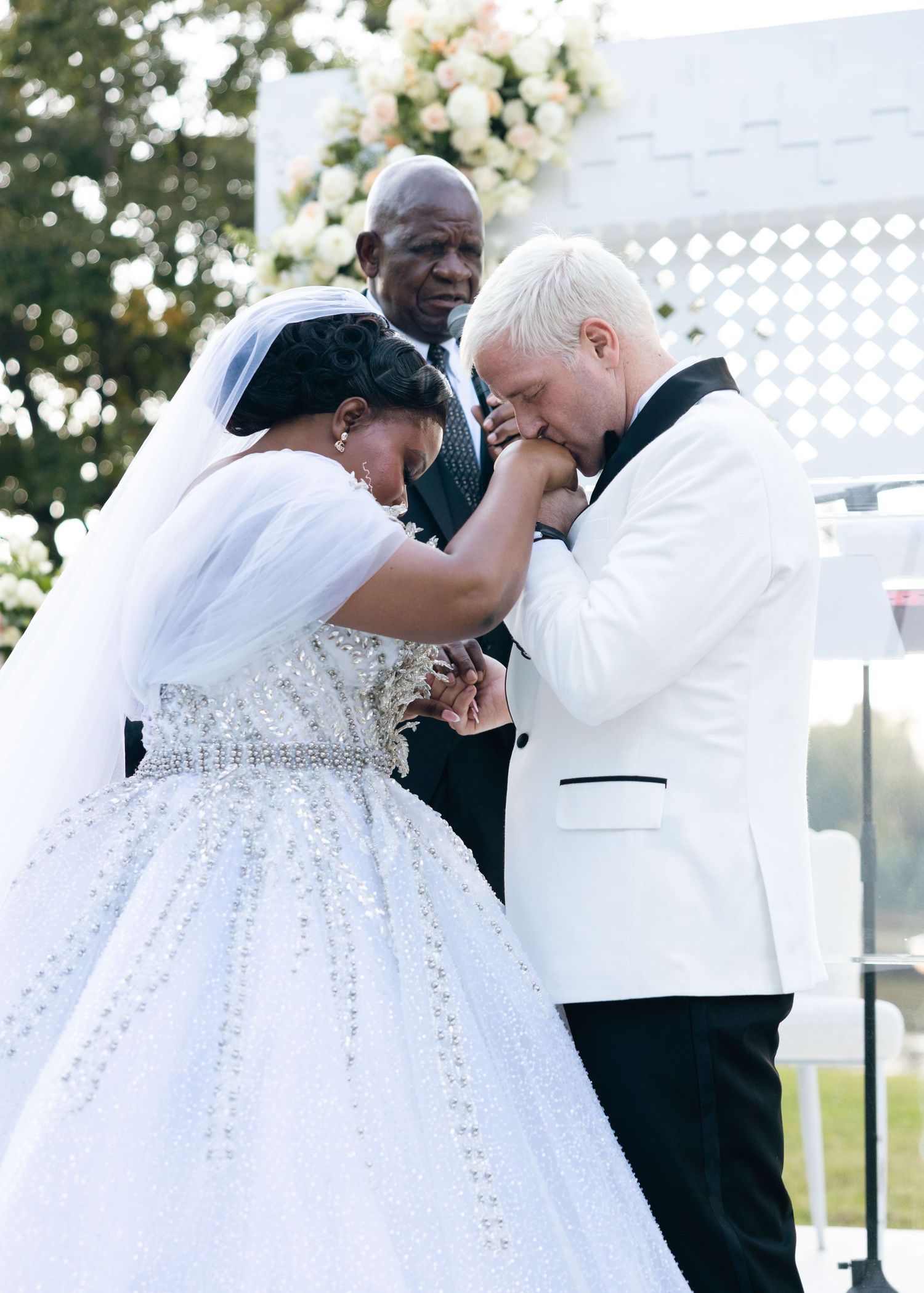 Groom in white tuxedo emotionally kisses the bride's hand during their outdoor wedding ceremony.