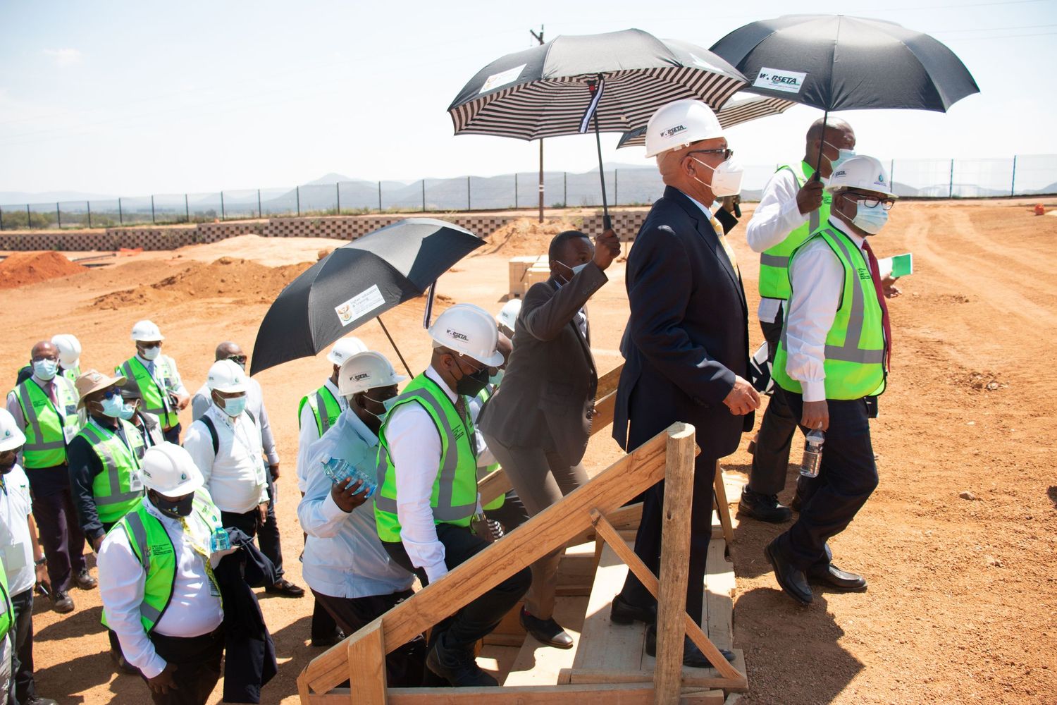 A group of officials wearing hard hats and safety vests walk up steps at an outdoor construction site.