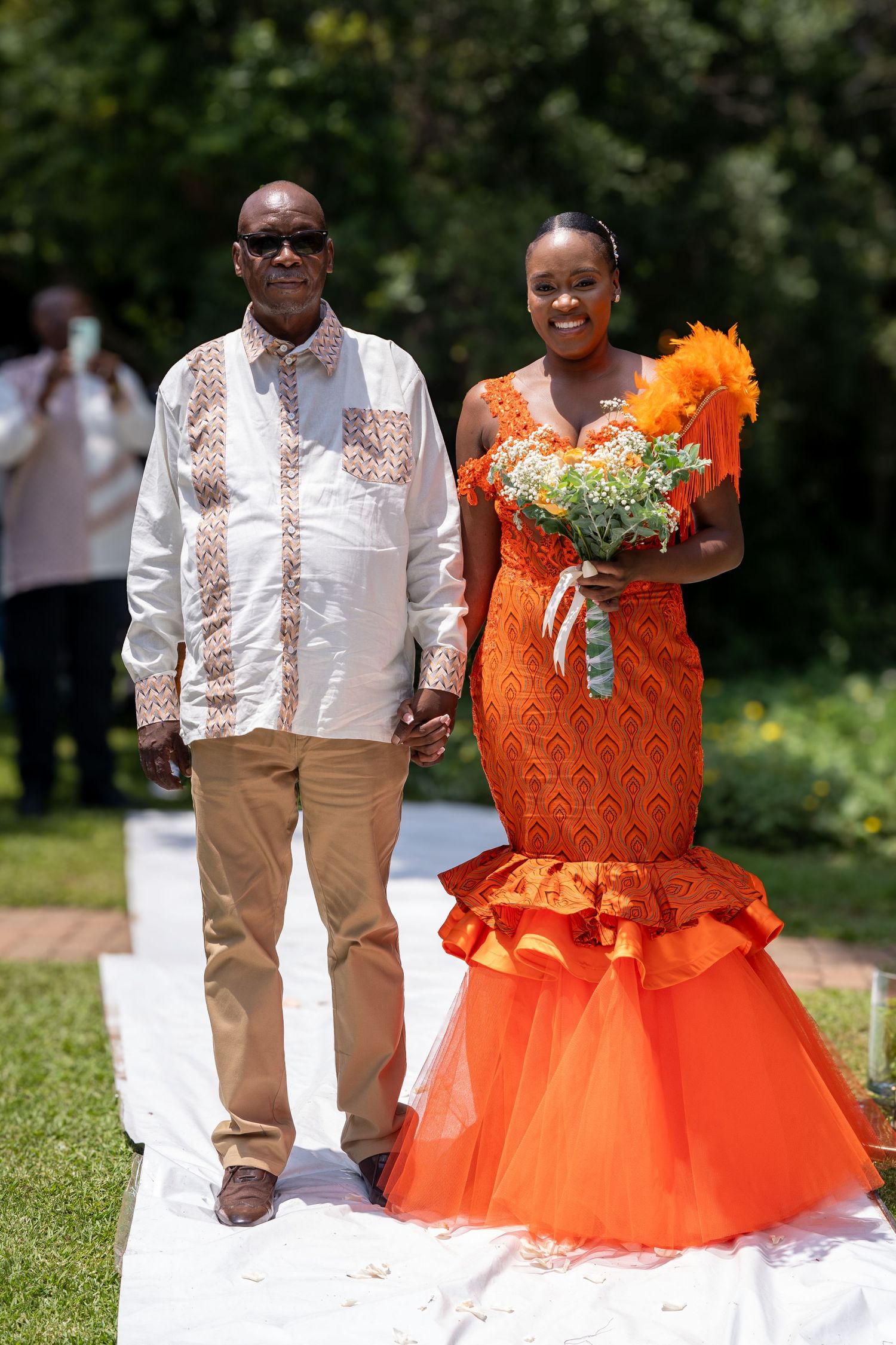 A bride in a stunning orange African print mermaid gown walks the aisle with an older gentleman at an outdoor wedding.