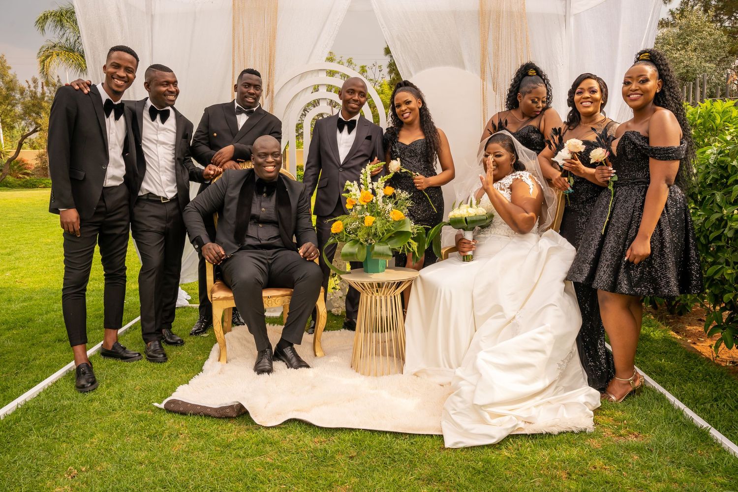 Wedding party in black attire posing with bride and groom at outdoor ceremony with floral backdrop.