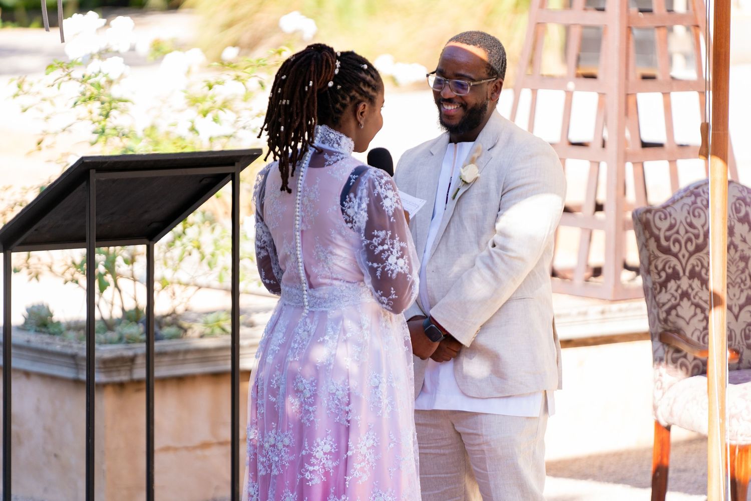 Groom in beige suit holds hands with bride in pink lace gown during intimate outdoor wedding vows.