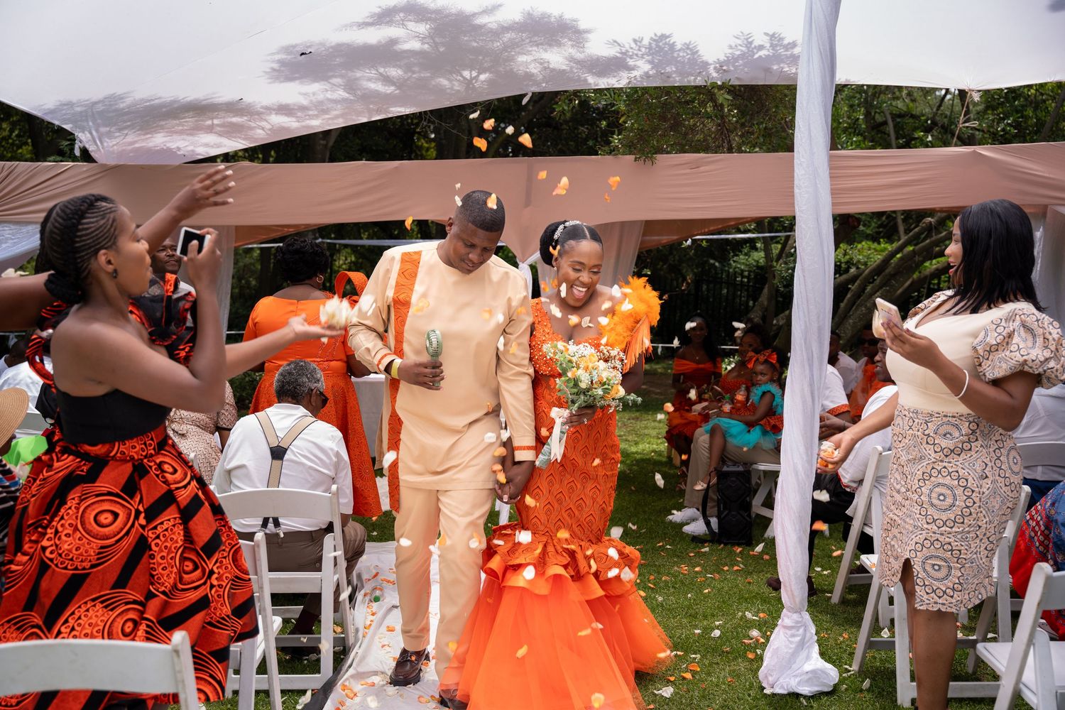 Couple in orange and beige traditional African attire walking down outdoor wedding ceremony aisle.