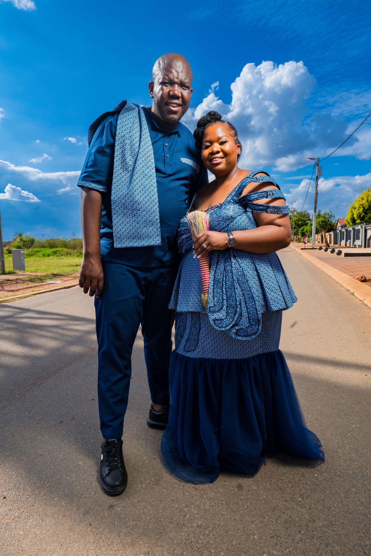 Couple in matching blue traditional African attire posing on a sunny street with blue sky backdrop.