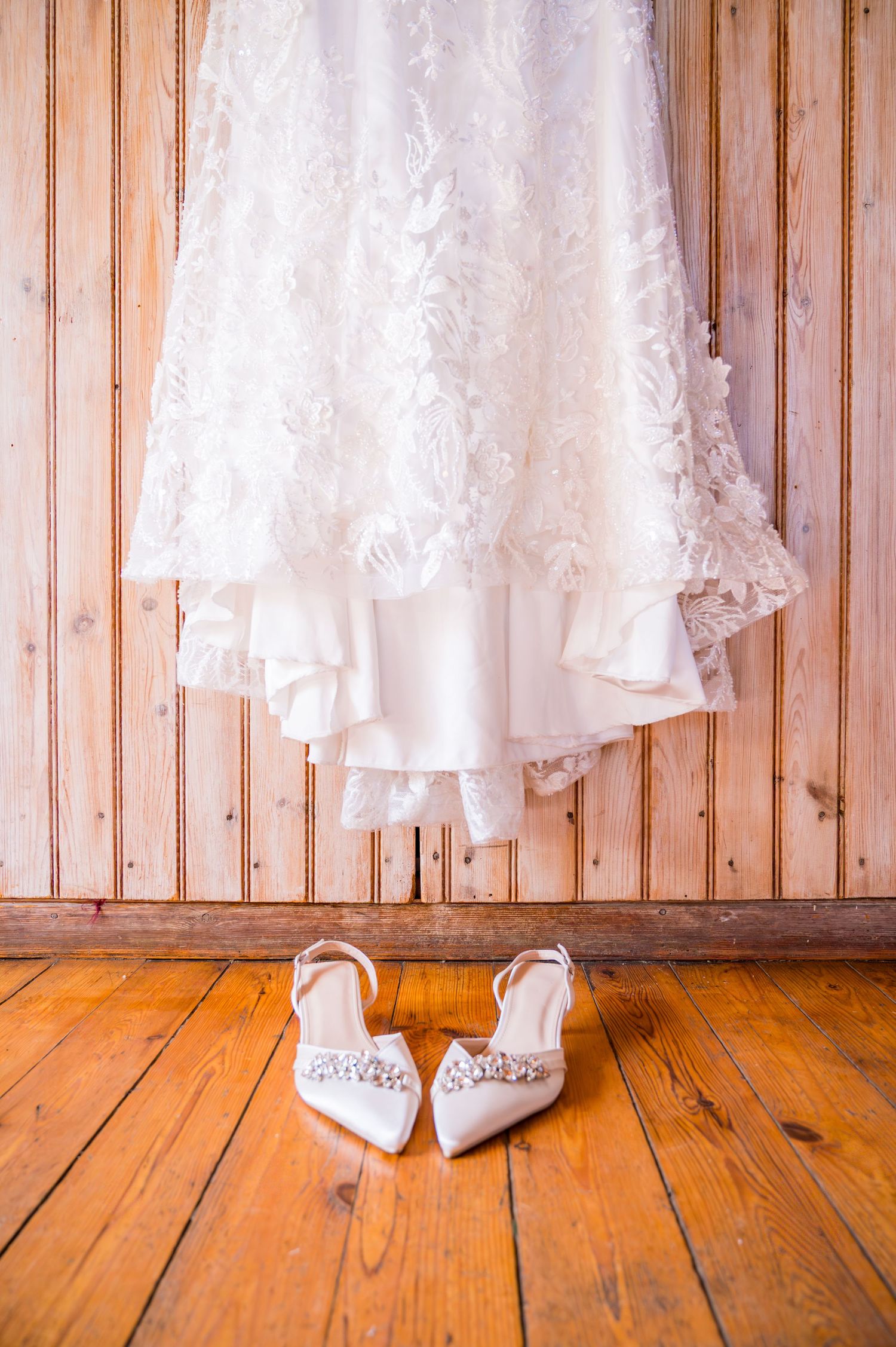 White lace wedding dress hanging above crystal-embellished bridal heels on rustic wooden floor.