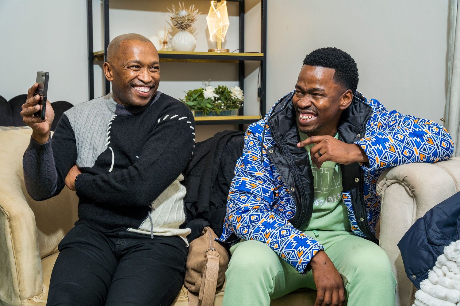 Two men laugh together on a couch at a social gathering, one wearing a vibrant blue patterned jacket.