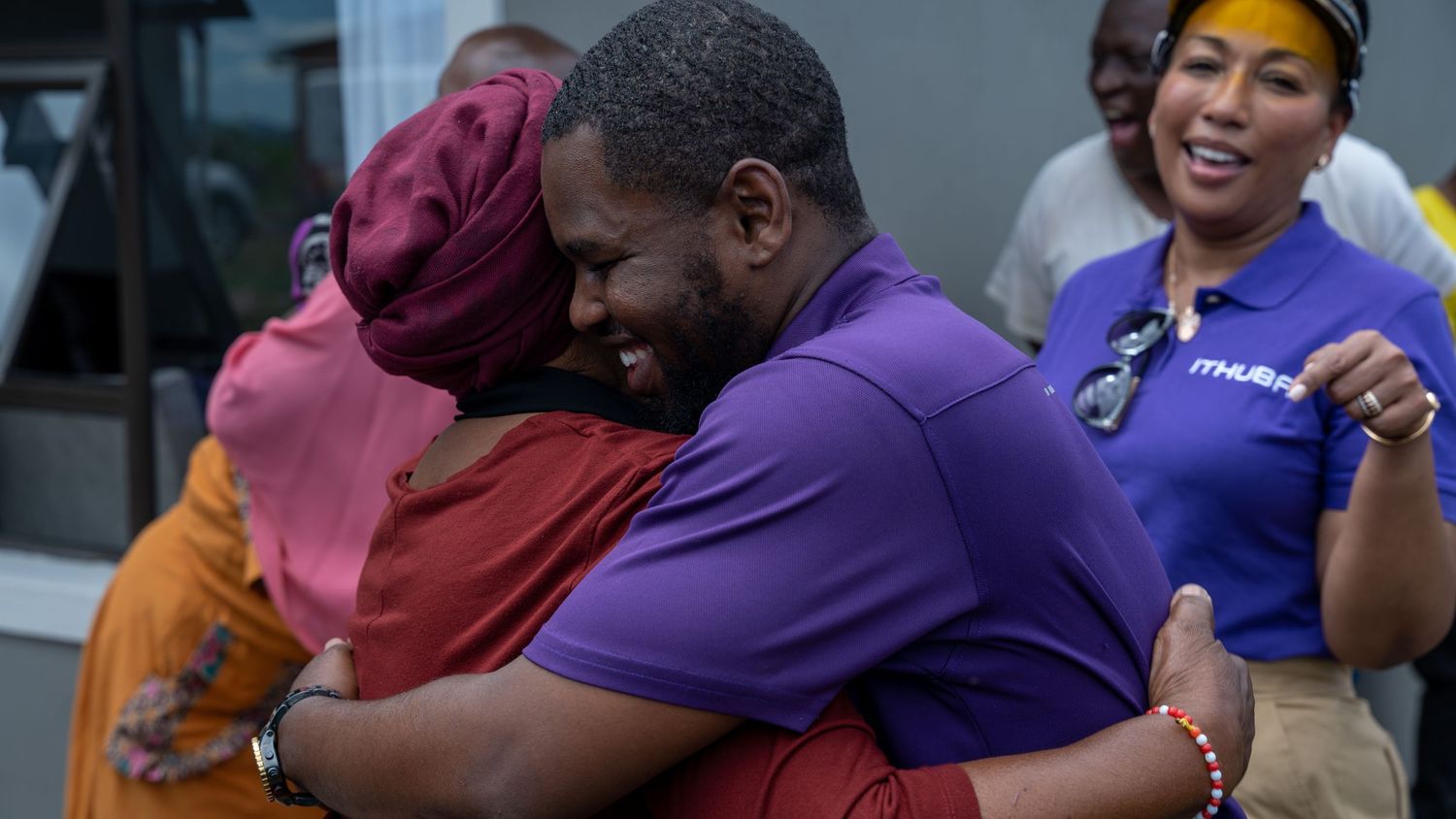 A joyful man in a purple shirt embraces a woman in a warm celebratory hug outdoors.