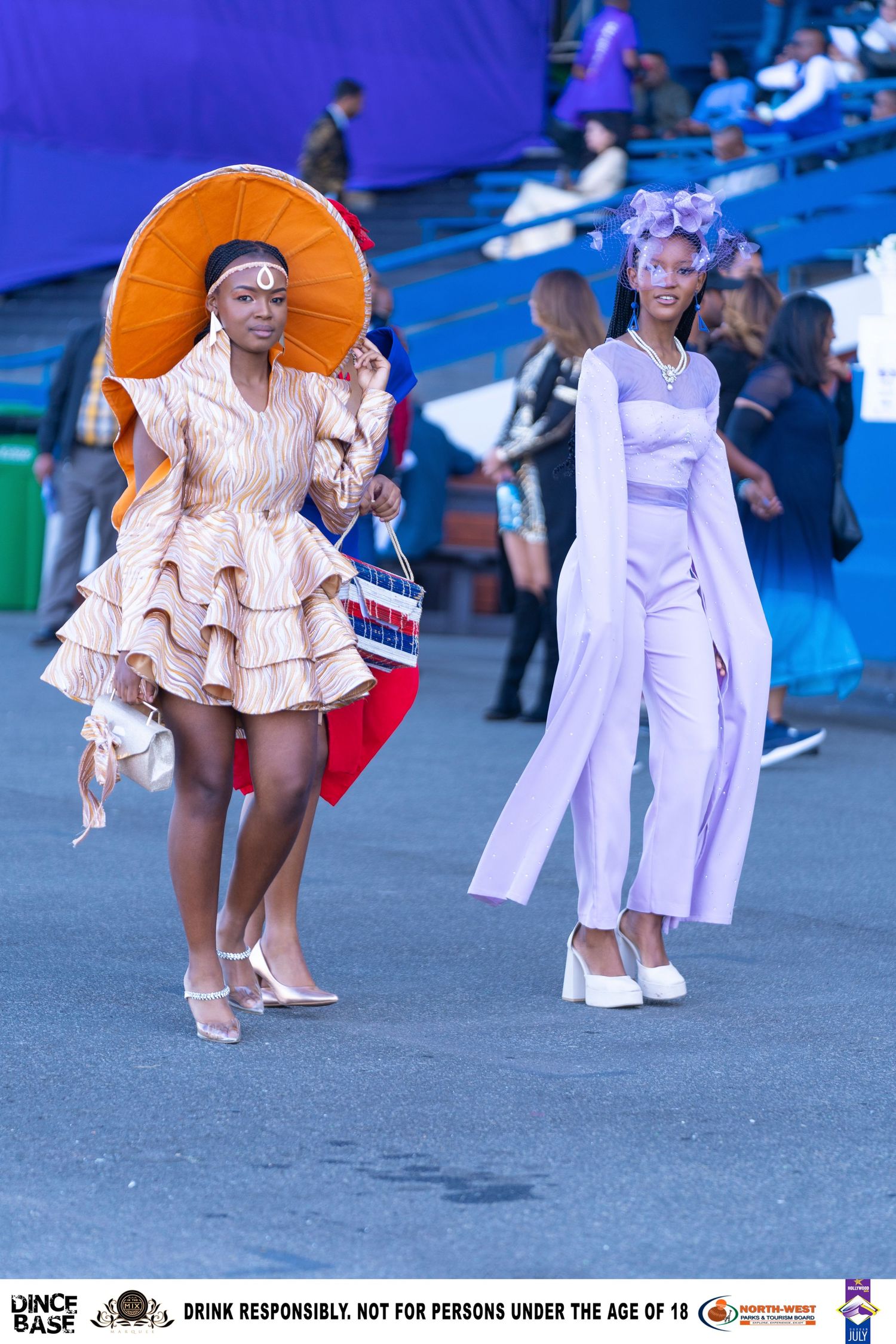 Two women in avant-garde fashion outfits with statement hats walking at an outdoor event.