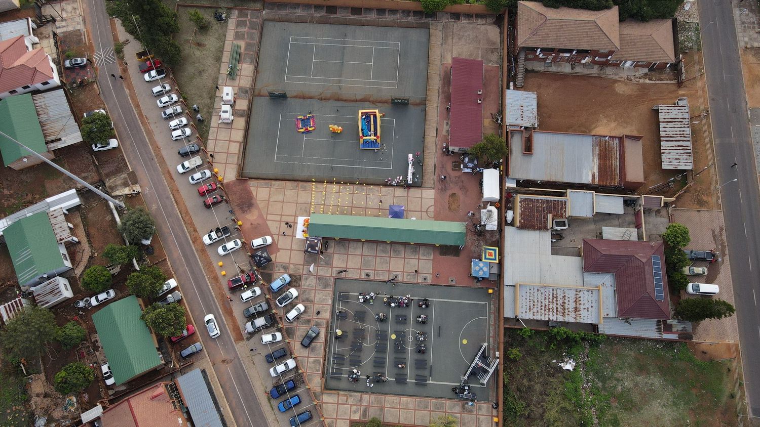 Aerial drone view of a tennis court, parking lot, and surrounding buildings in an urban African neighborhood.