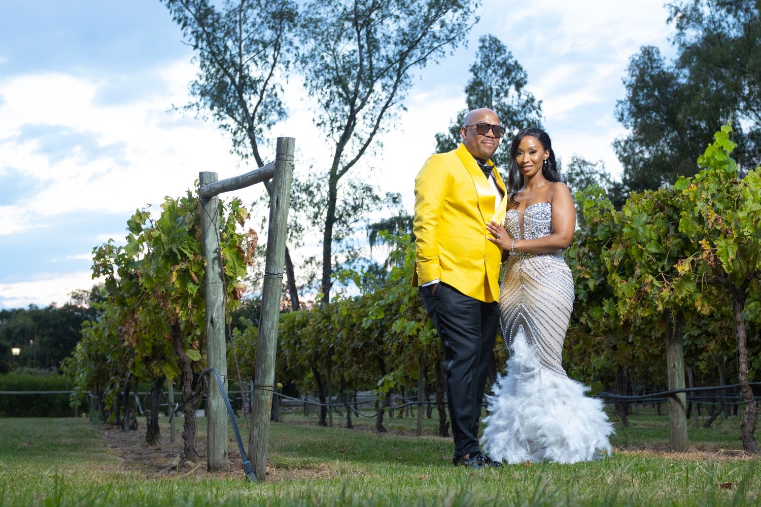 Couple poses in vineyard, groom in yellow suit and bride in silver gown at sunset.