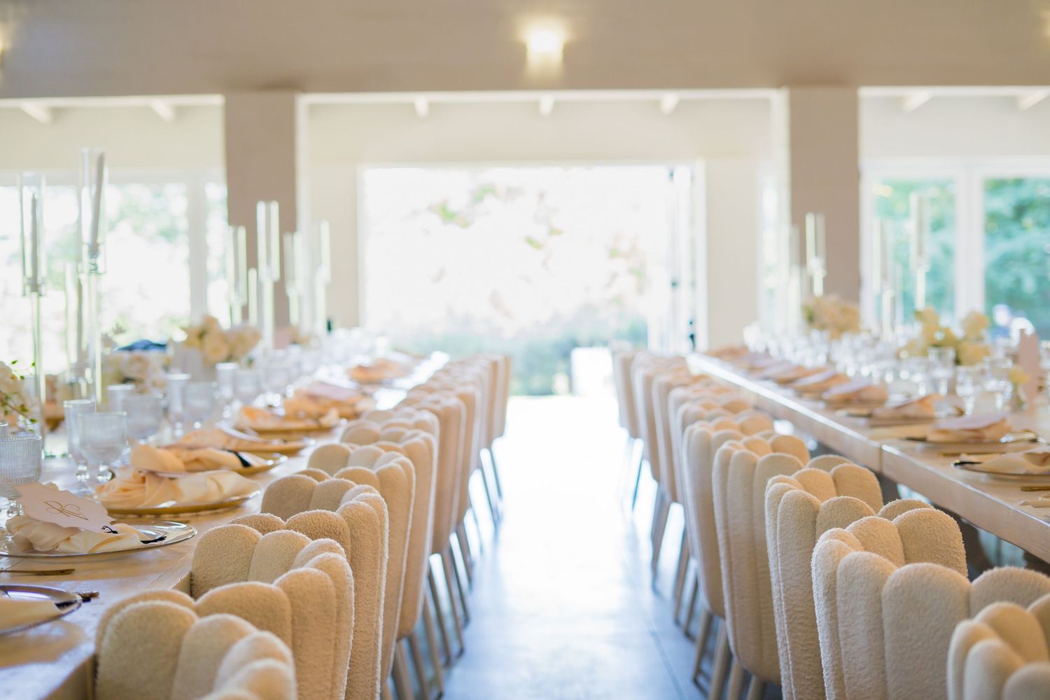 Elegant wedding reception hall with long tables, white chairs, gold place settings, and tall candles.