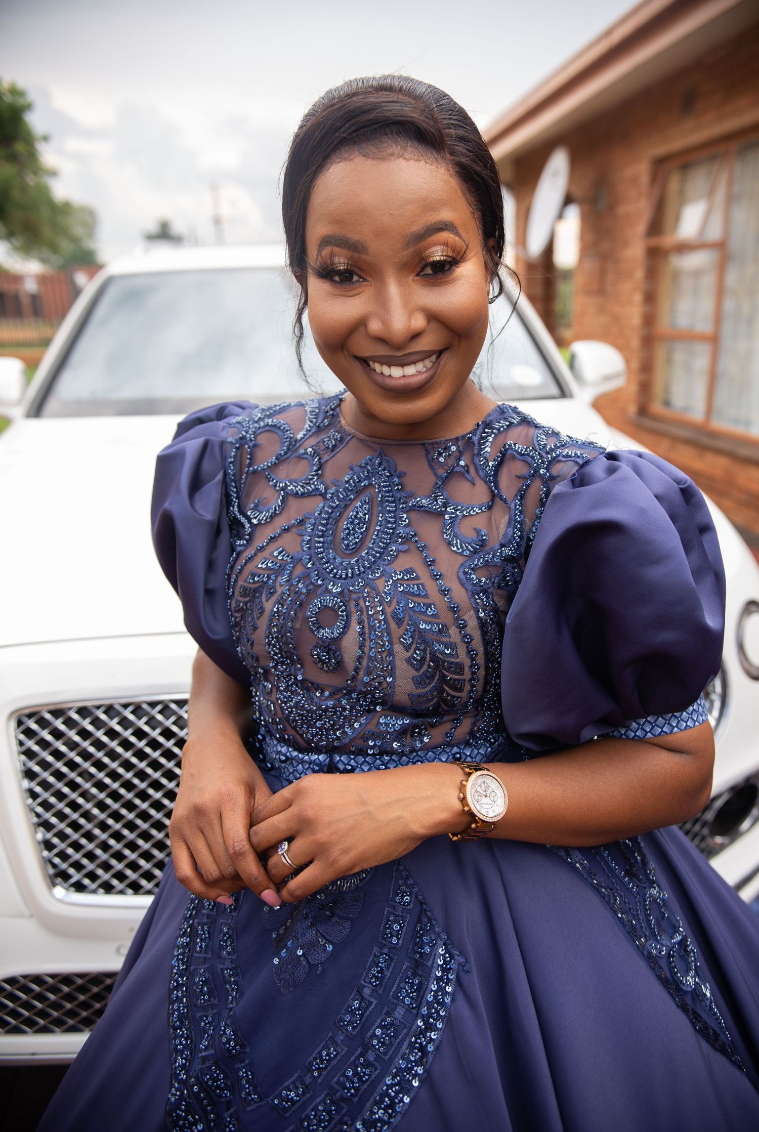 Woman in elegant navy blue beaded ball gown with sheer bodice and puff sleeves smiling at outdoor event.