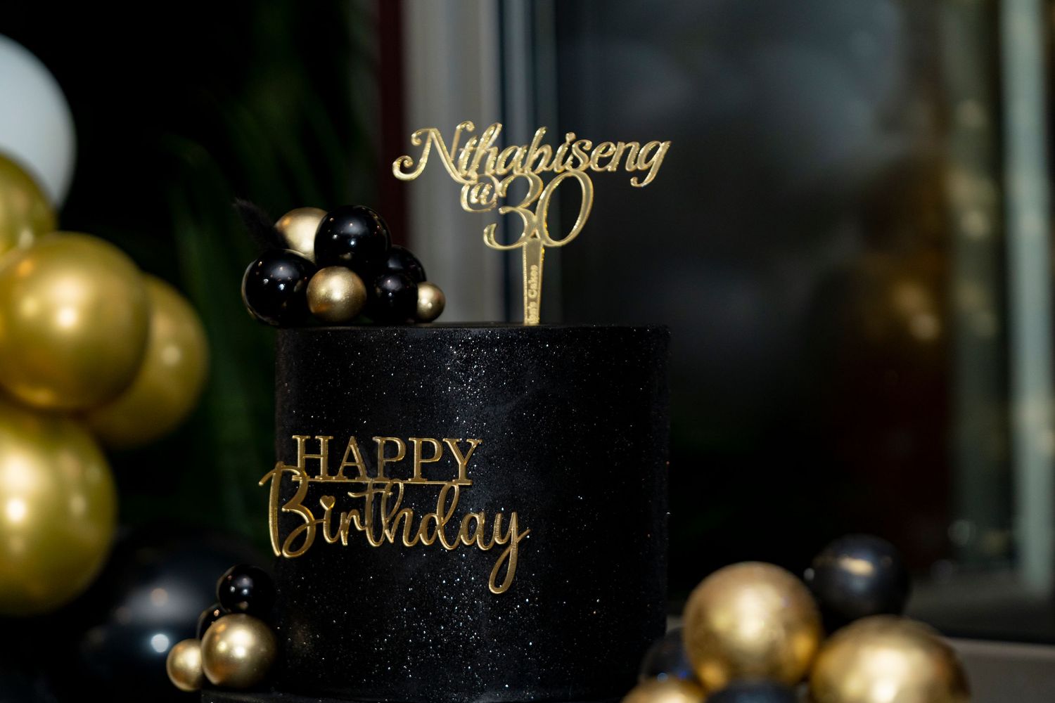 Black and gold Happy Birthday cake with Nthabilseng turns 30 topper and balloons.