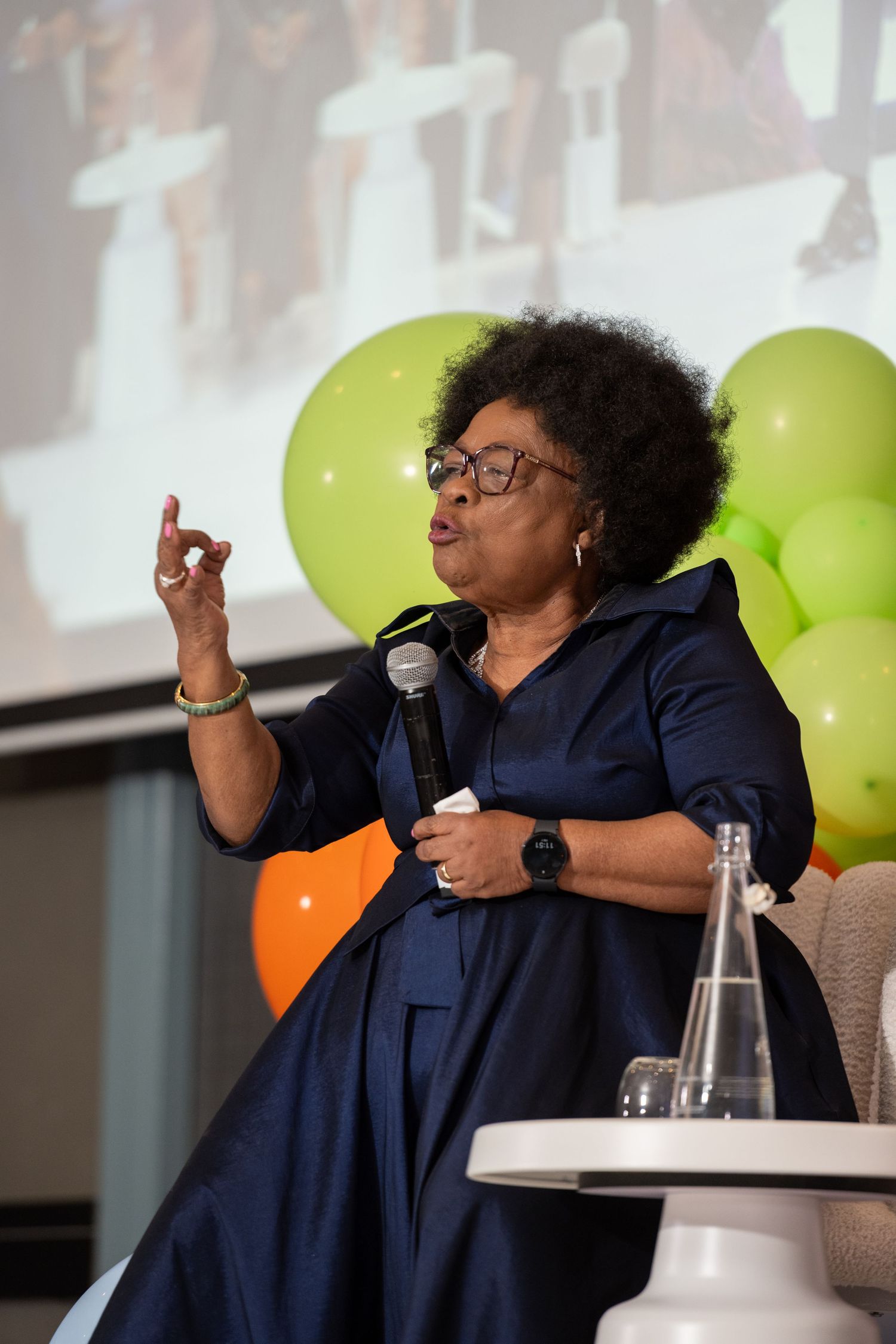 Woman in navy dress speaking at event with microphone, gesturing with finger raised, colorful balloons in background.