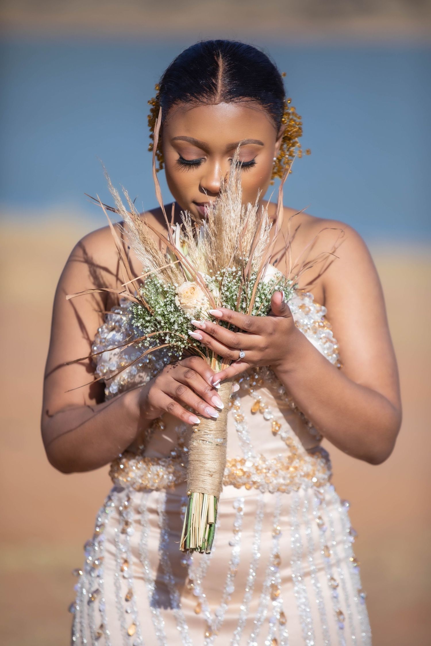 Beautiful African bride in beaded gown holding rustic dried flower bouquet outdoors near water.