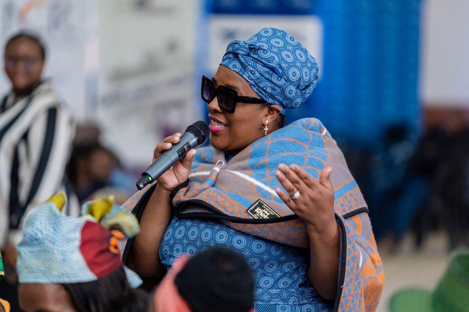 Woman in blue traditional attire and headwrap speaks into microphone at outdoor community event.