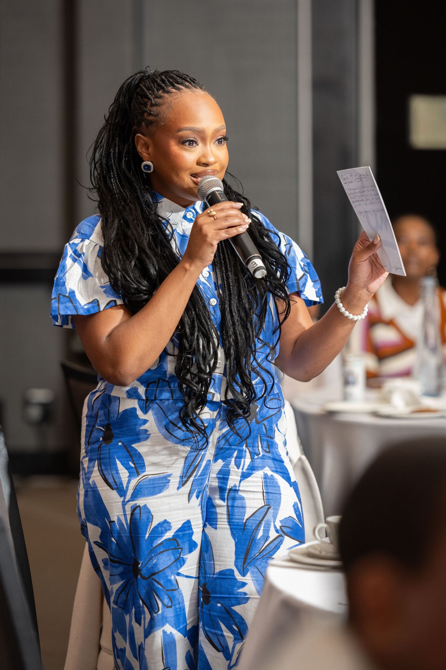 Woman in blue floral dress speaks at microphone holding notes at elegant indoor event.