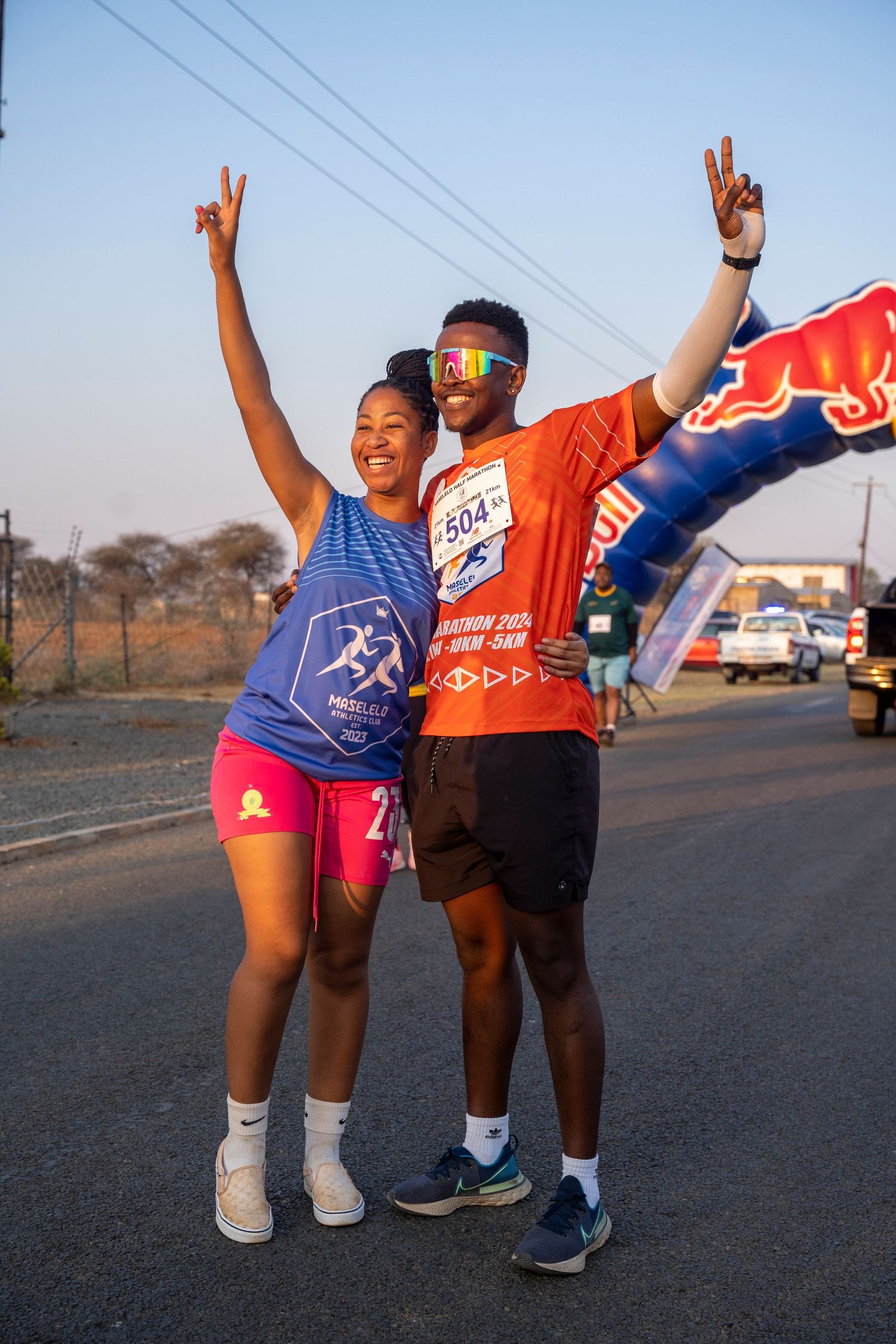 Two runners celebrate with peace signs at a Red Bull marathon finish line at sunrise.