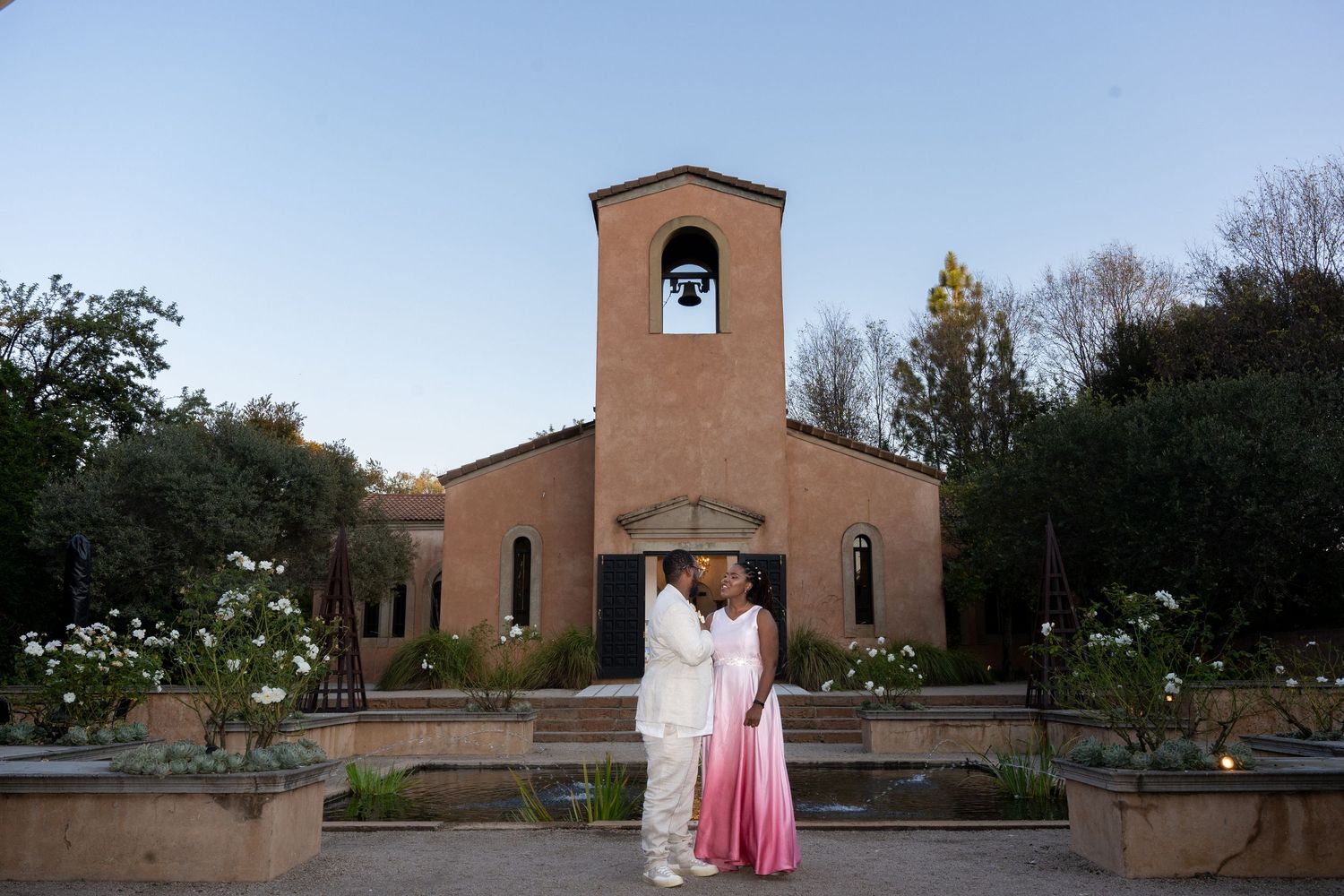 Newlyweds embracing in front of a rustic chapel at dusk in a romantic outdoor wedding setting.