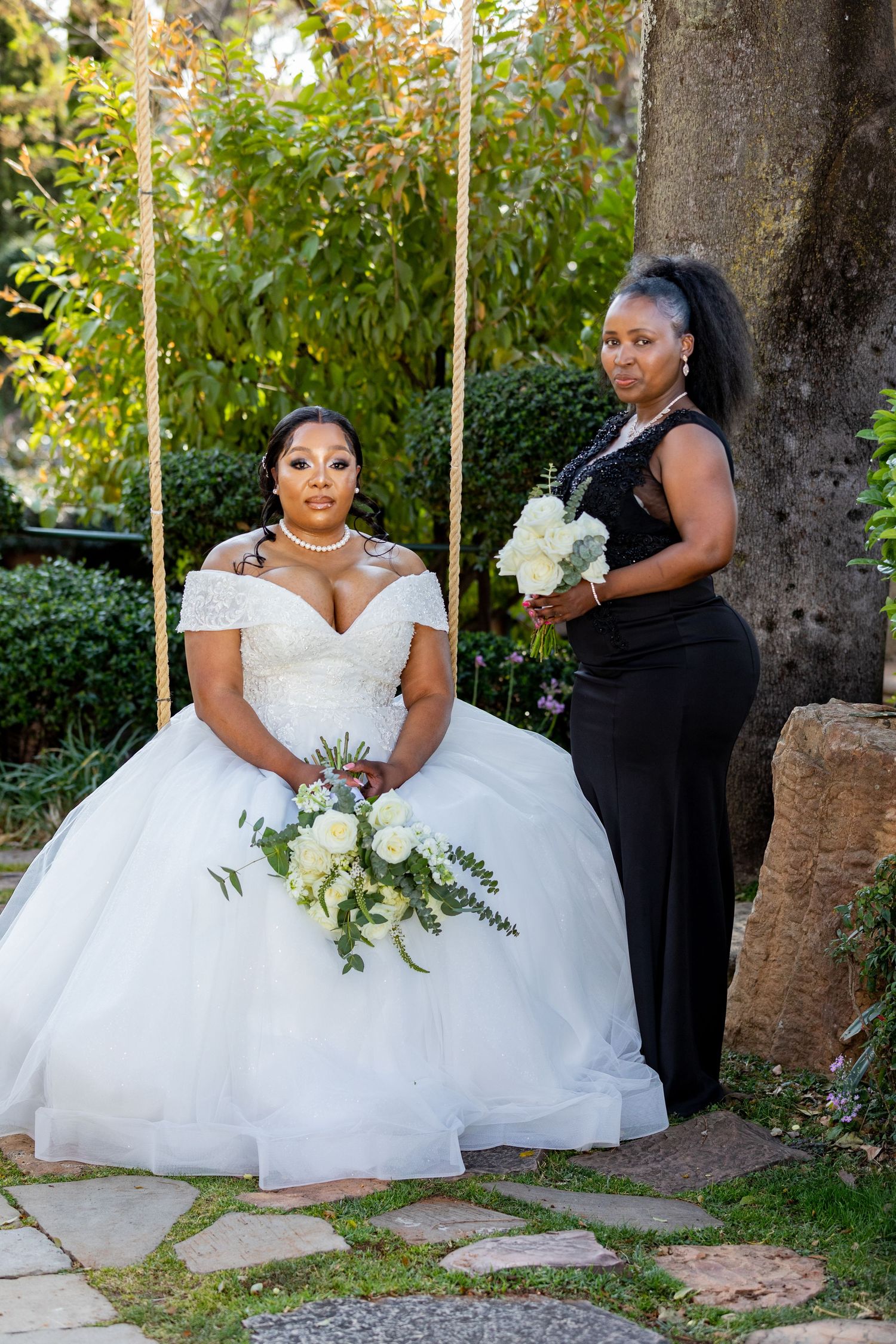 Bride in white ball gown seated on swing with bridesmaid in black dress holding white flowers.
