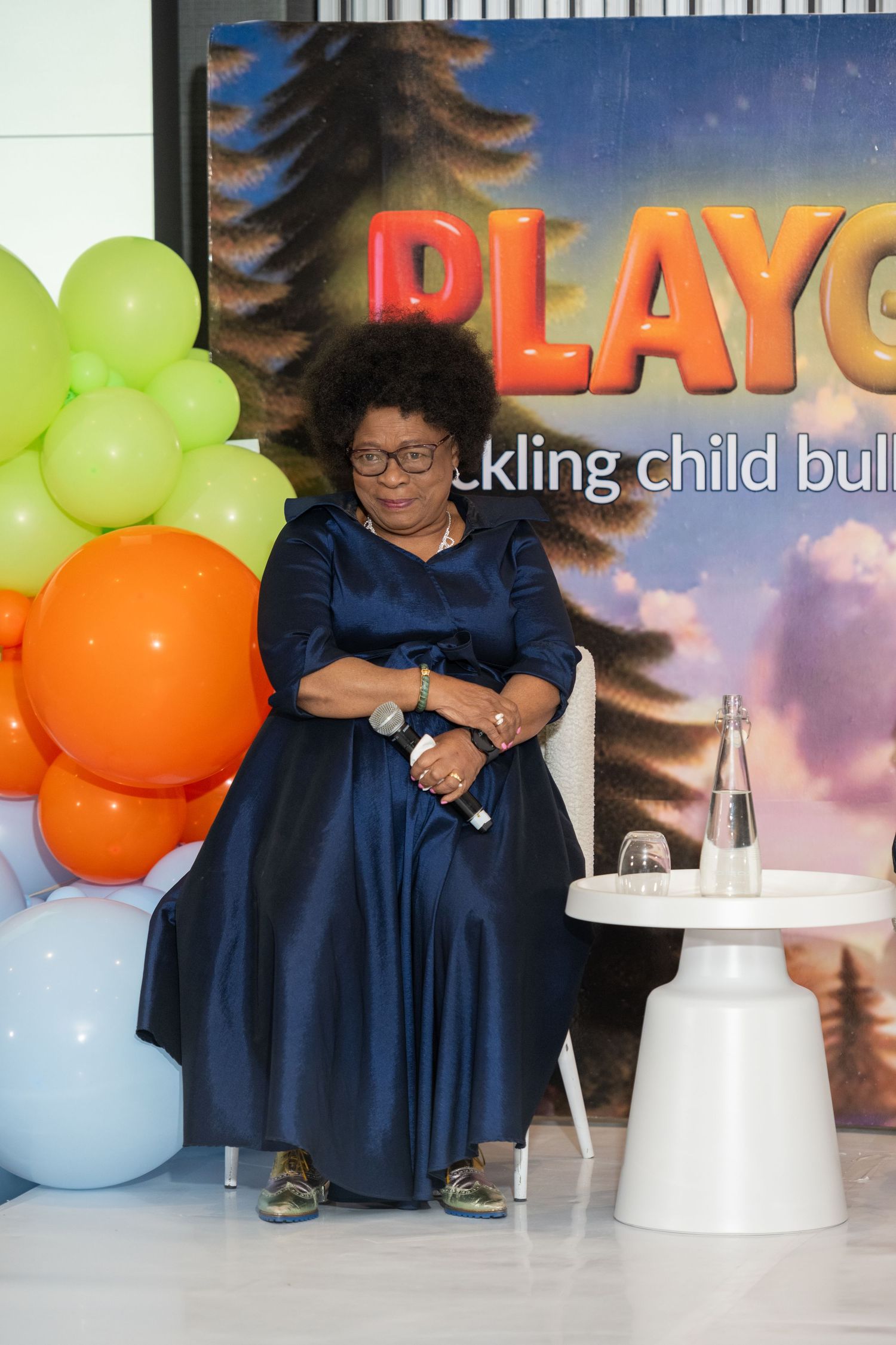 Woman in navy blue satin dress seated at Playgrounds child bullying awareness event.