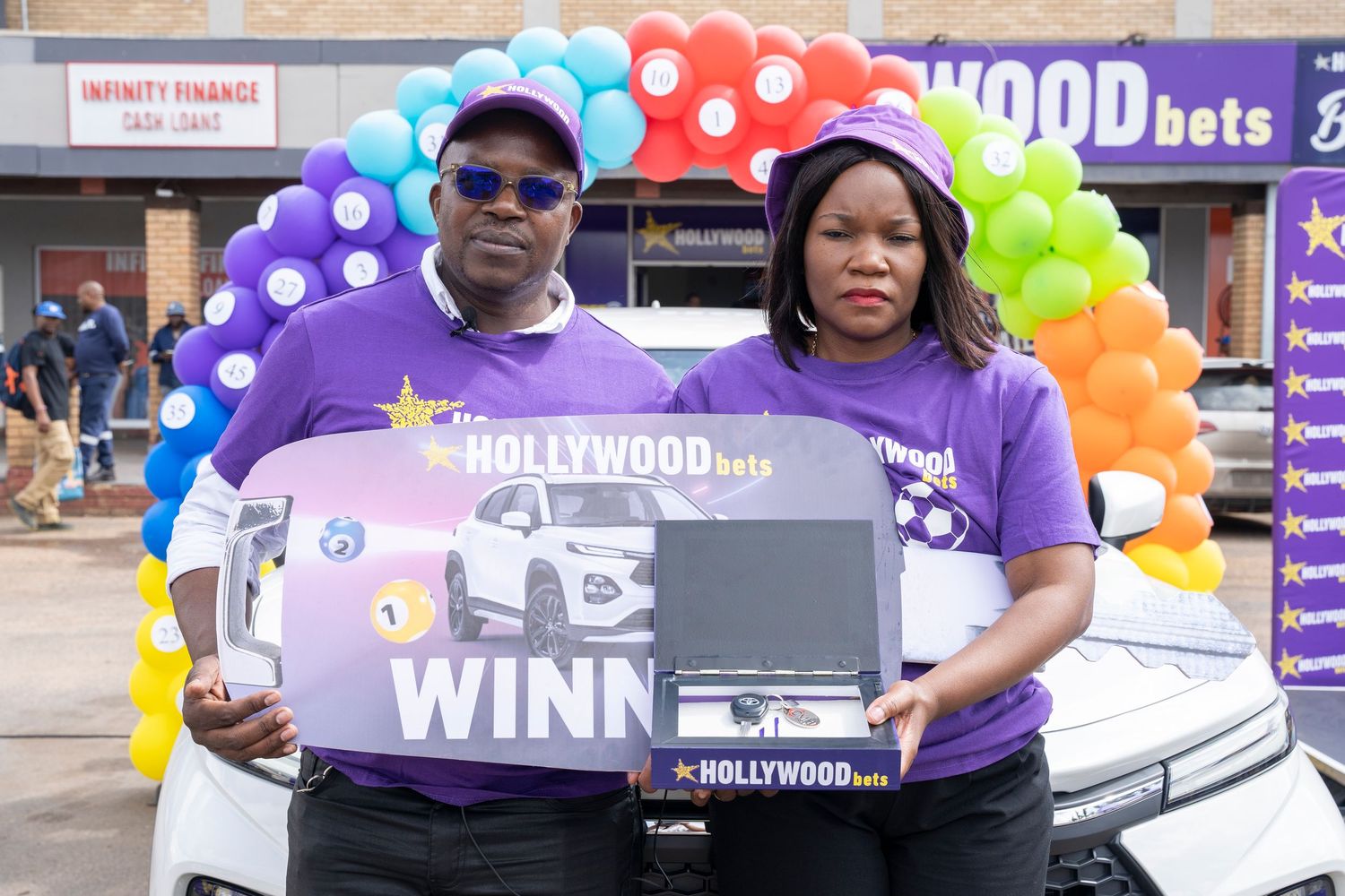Couple poses proudly with Hollywoodbets winner boards and prize in front of branded backdrop.