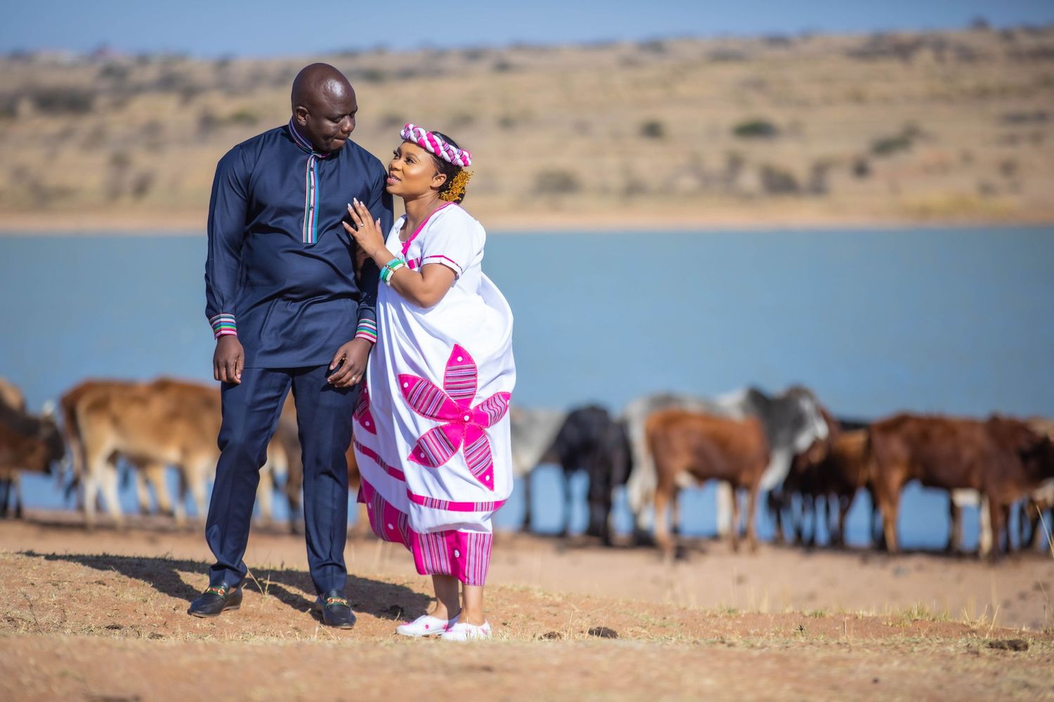African couple in traditional attire standing near a lake with cattle grazing in the background.