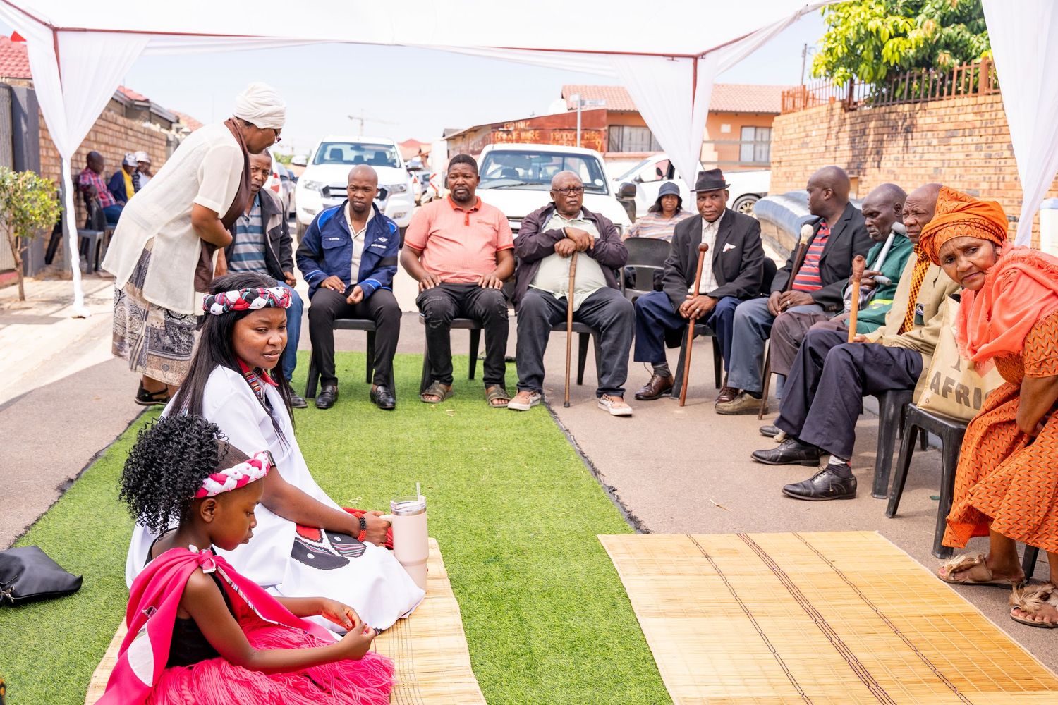 Traditional African cultural ceremony with young girl in white dress kneeling before seated elders on green grass outdoor setting.