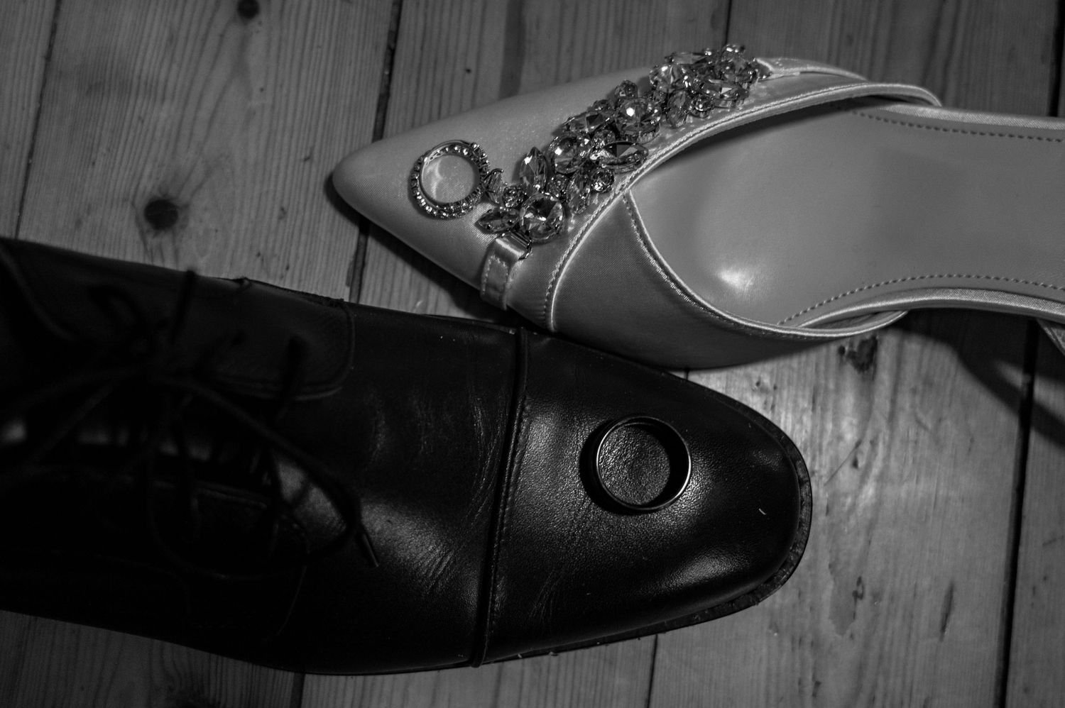 Black and white photo of bejeweled bridal heel beside grooms dress shoe on wood floor.