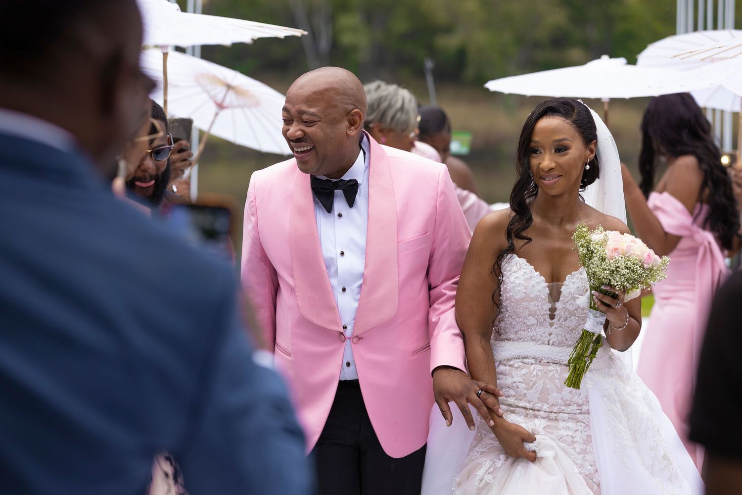 Smiling bride in lace gown walks with escort past guests at a beautiful outdoor wedding ceremony.