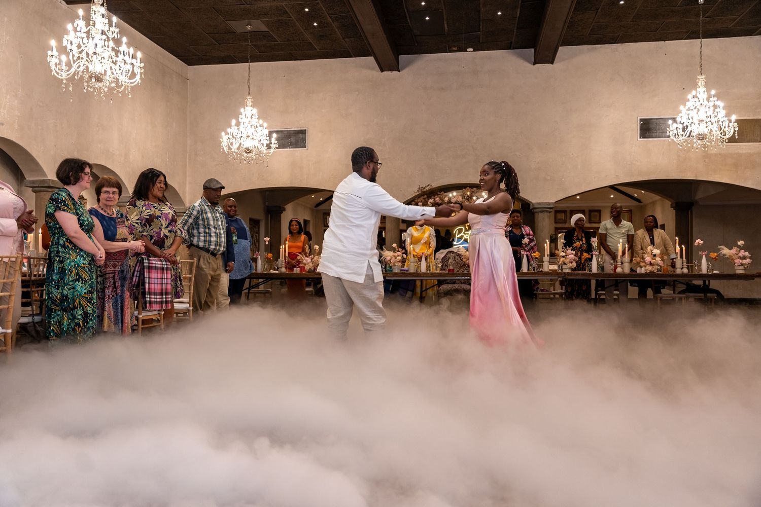 Newlyweds share first dance surrounded by guests in an elegant hall with dramatic fog effects.
