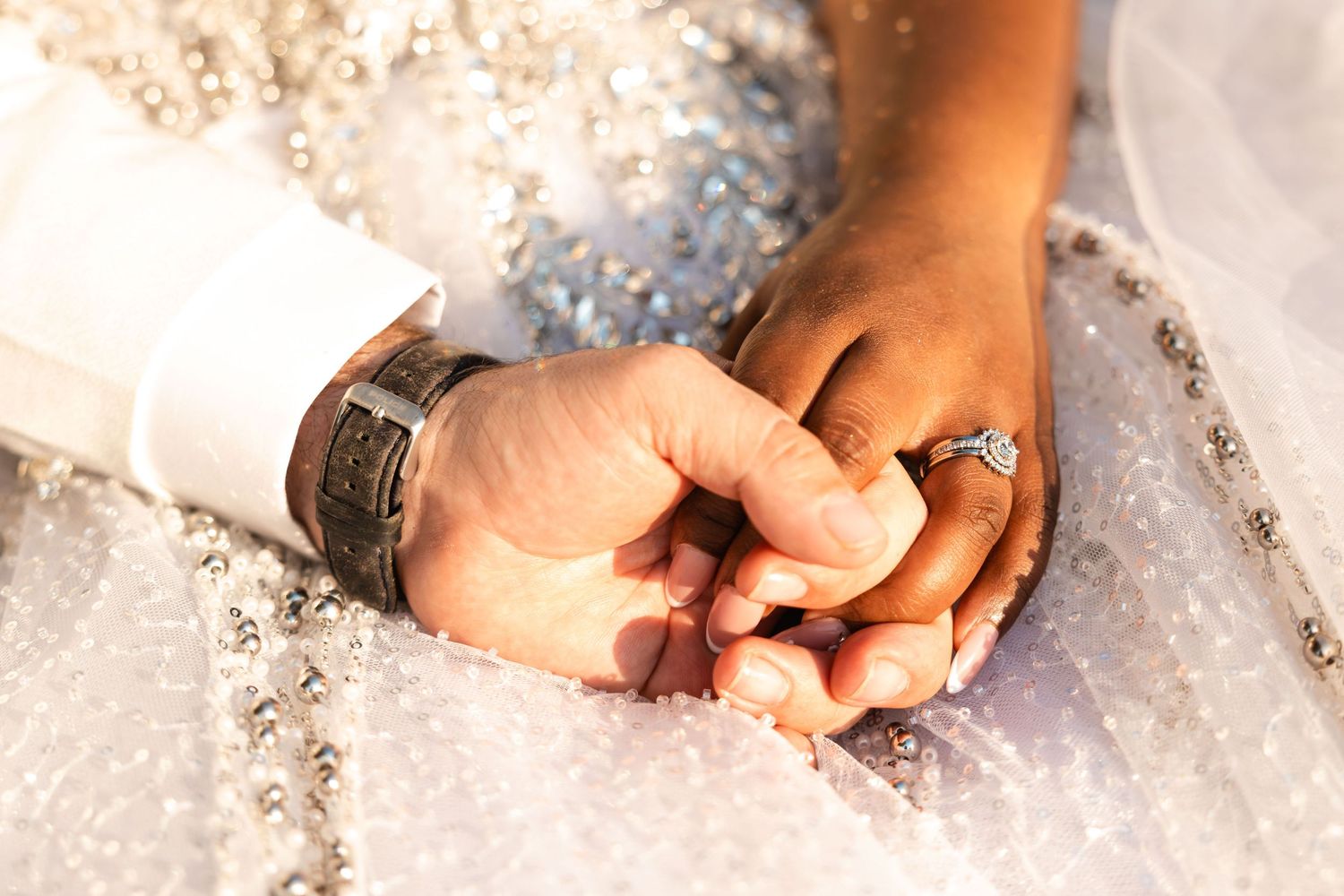 Close-up of interracial couple holding hands with diamond engagement ring and watch on wedding day.