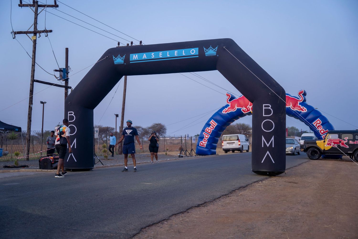 An inflatable Maselelo Boma and Red Bull branded finish line arch stands on a rural road at dusk.