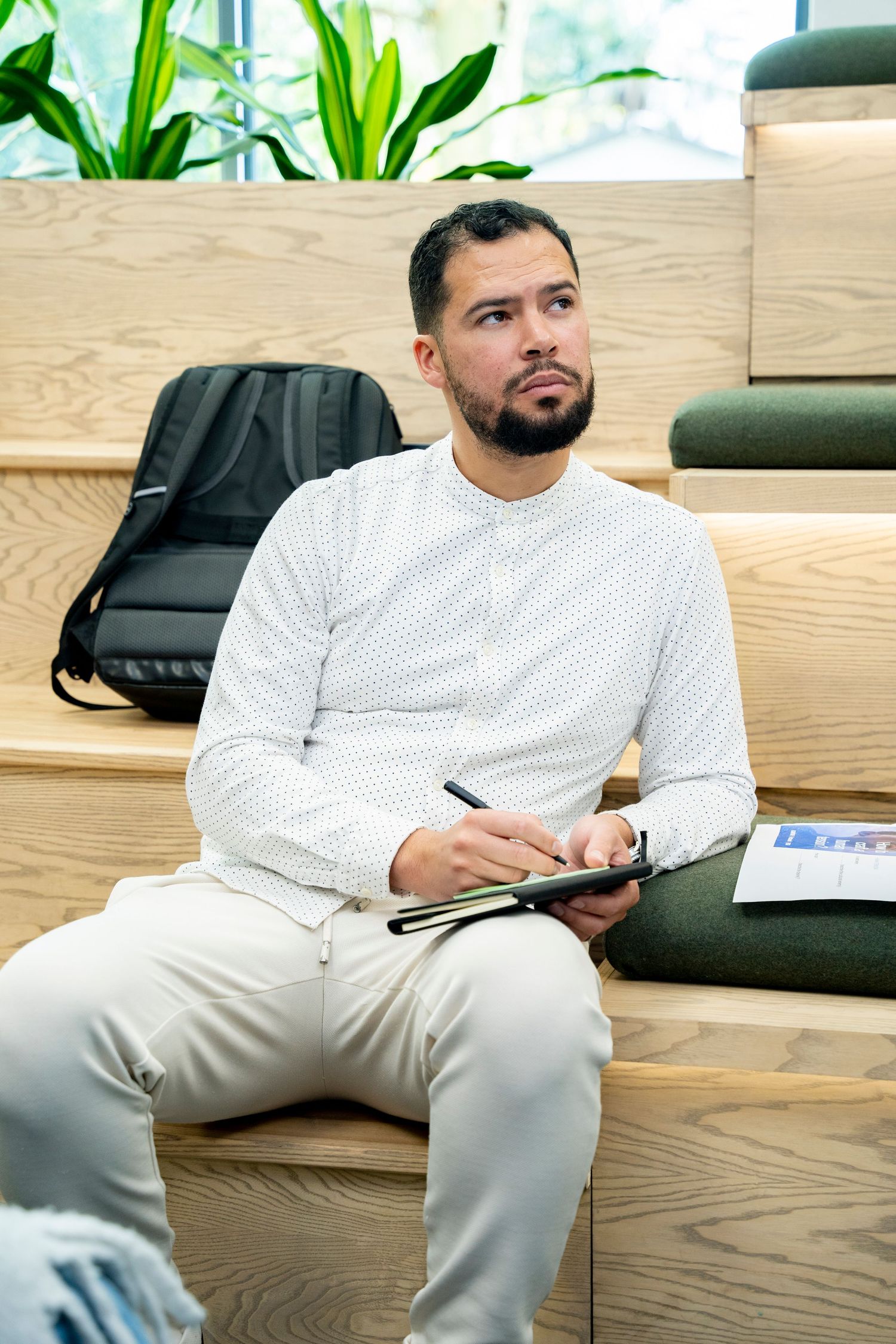 A bearded man in a white shirt sits on wooden steps, holding a pen and notebook, with a backpack beside him.
