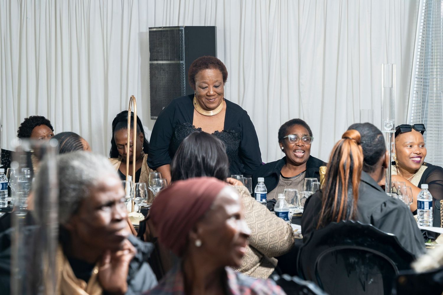 Elegantly dressed woman stands and engages with seated guests at a formal dinner gathering.