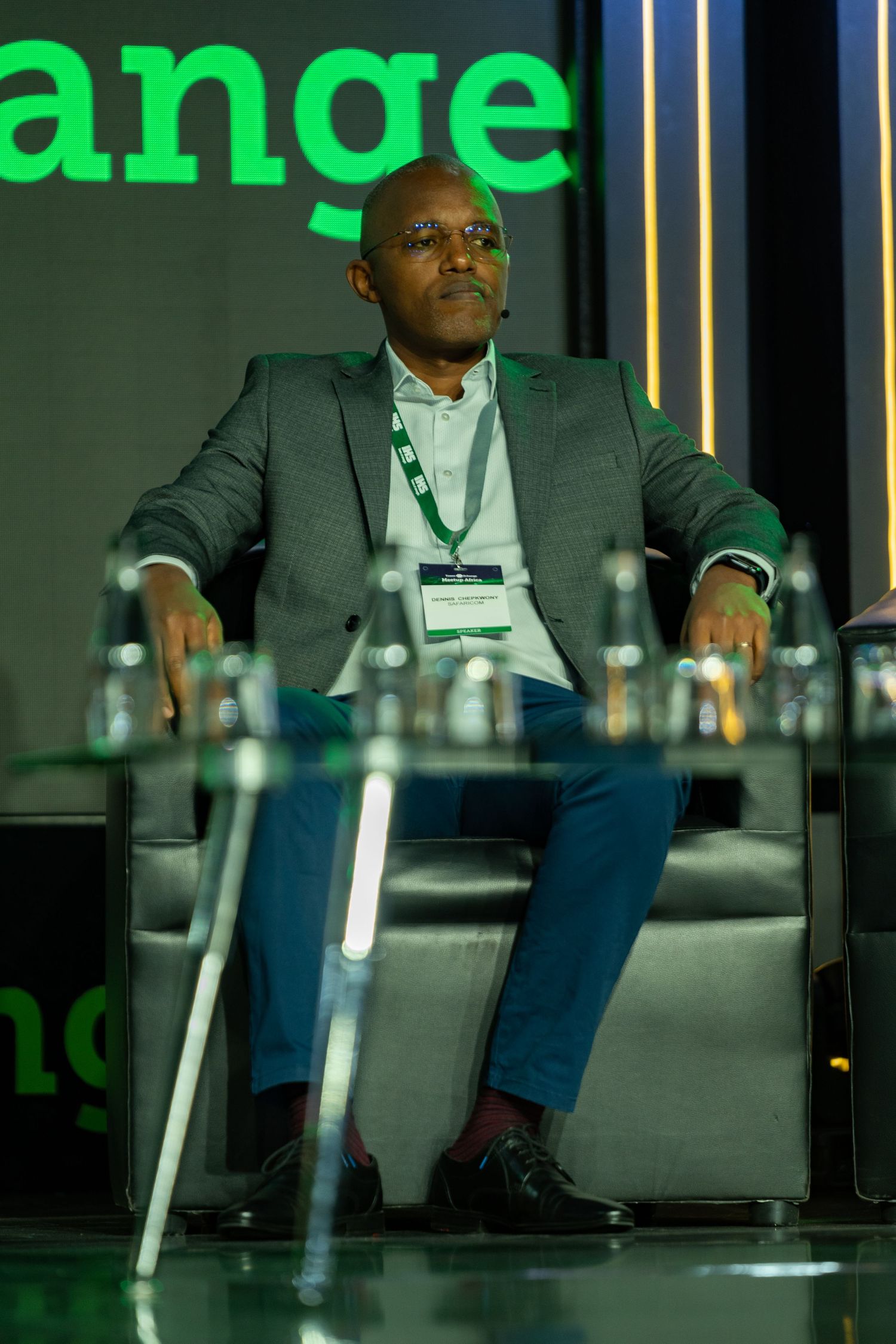 A man in a gray suit with a lanyard sits attentively in a chair at a professional conference event.