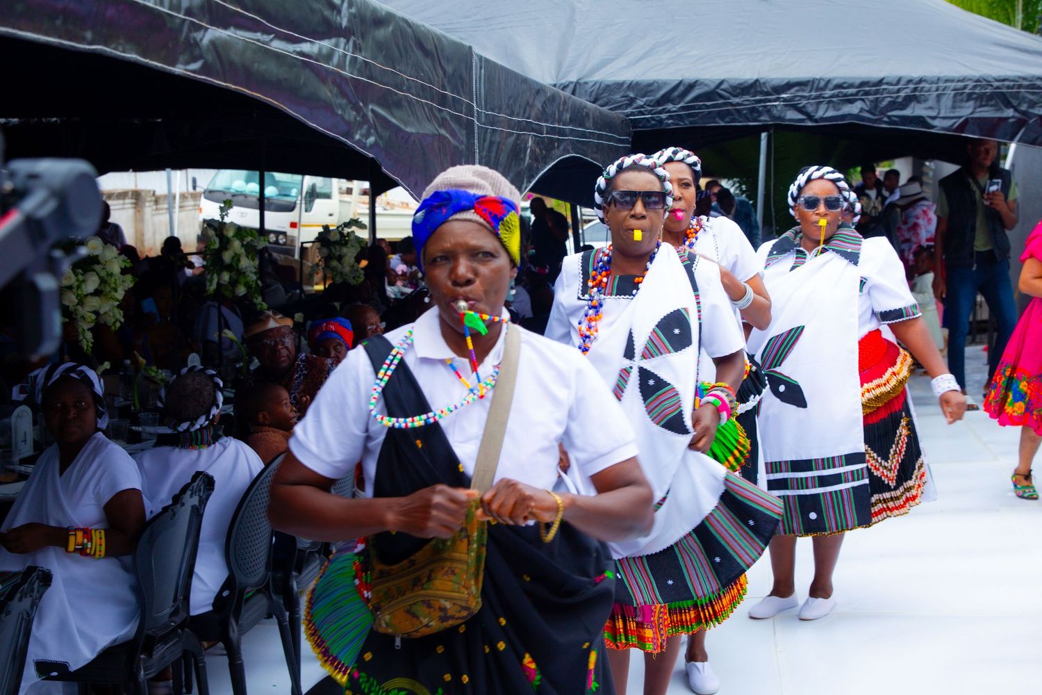 Traditional African ceremony with participants in Xhosa attire walking under a white tent.