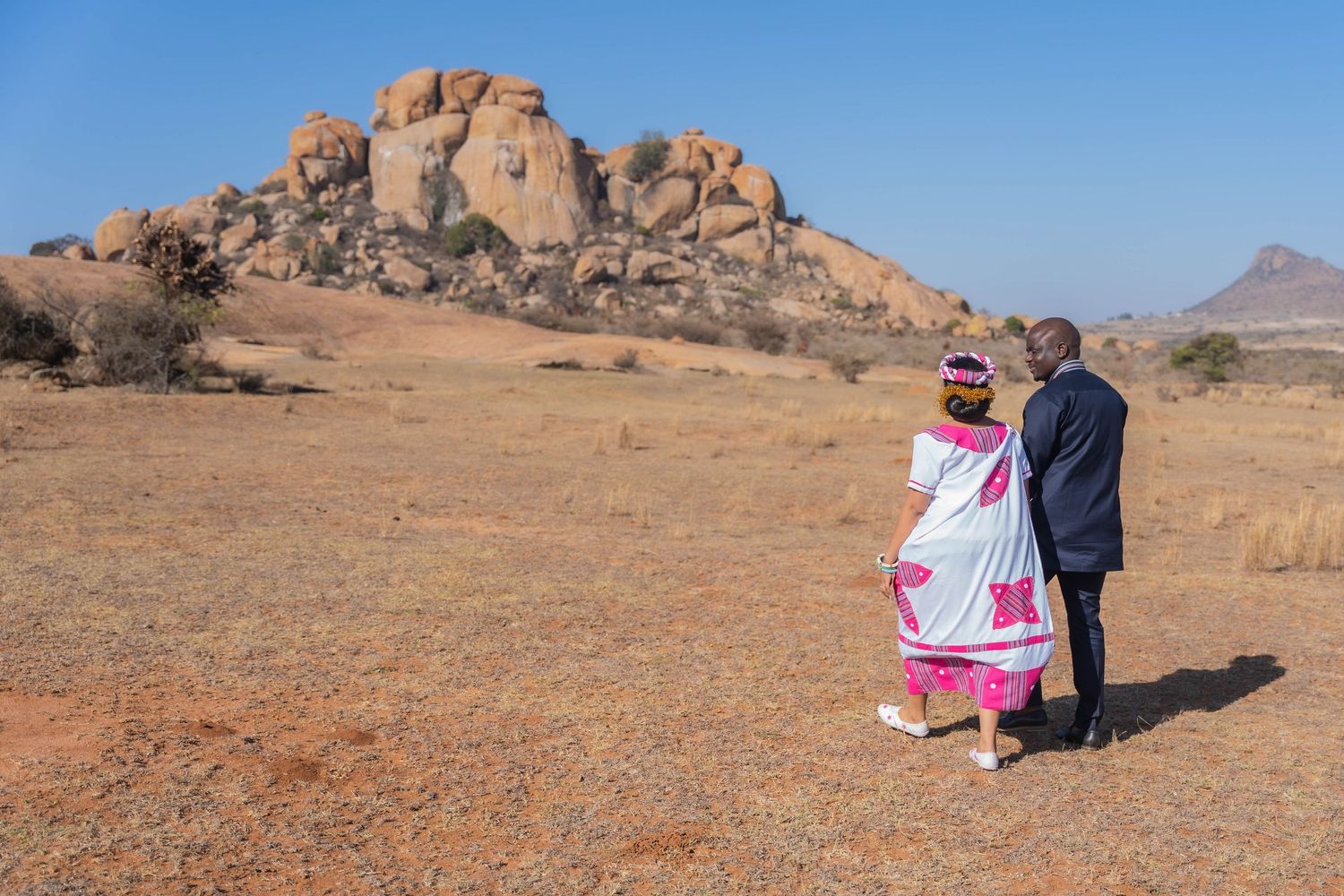 African couple in traditional and formal attire standing together in an arid landscape with rocky formations.