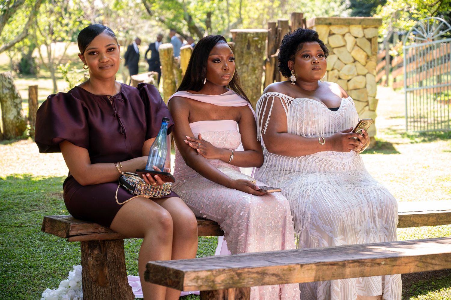 Three elegantly dressed women sit on a bench at an outdoor wedding ceremony.