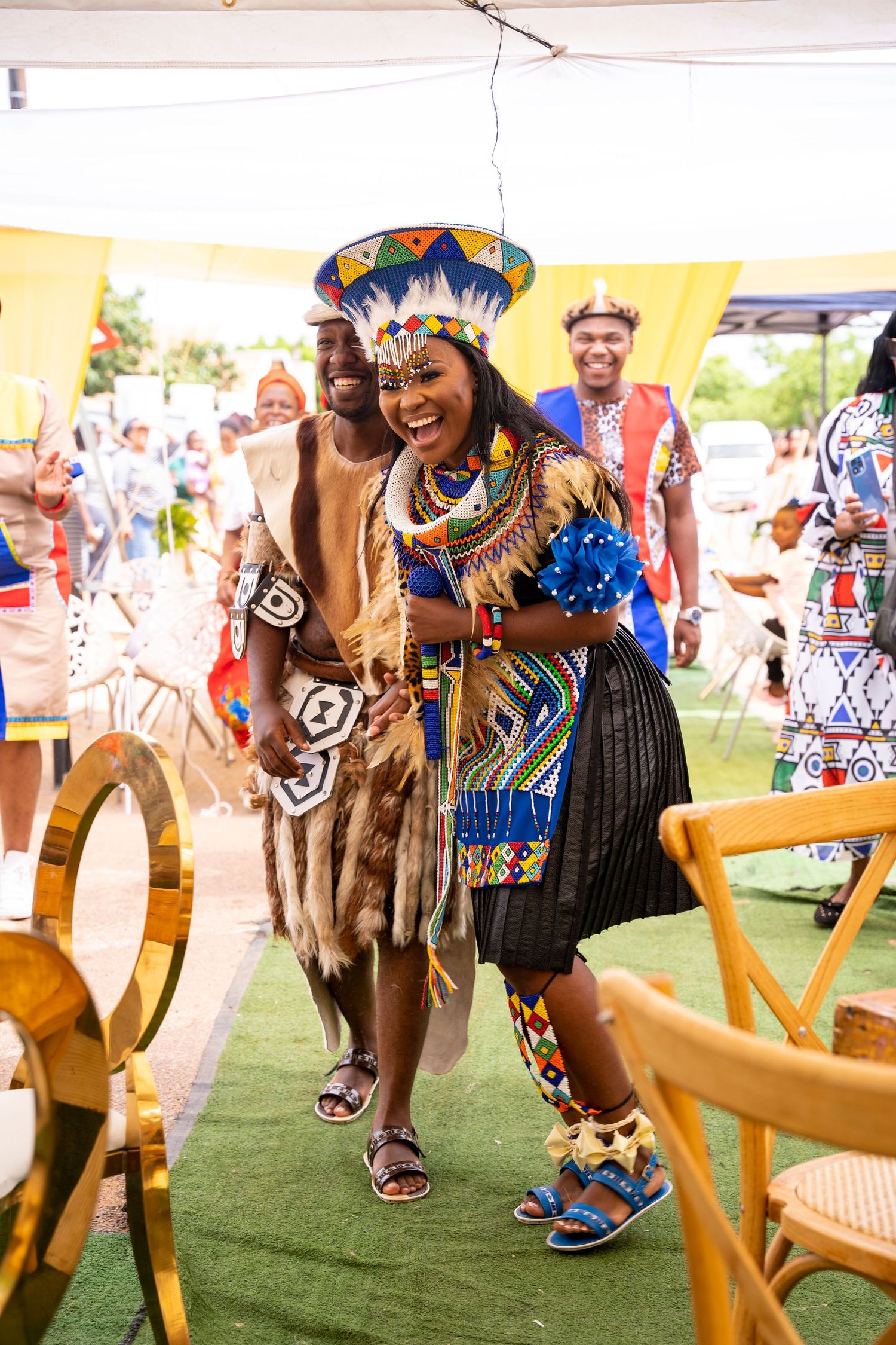 Laughing Zulu bride and groom in elaborate traditional beaded costumes walk together through decorated wedding venue.