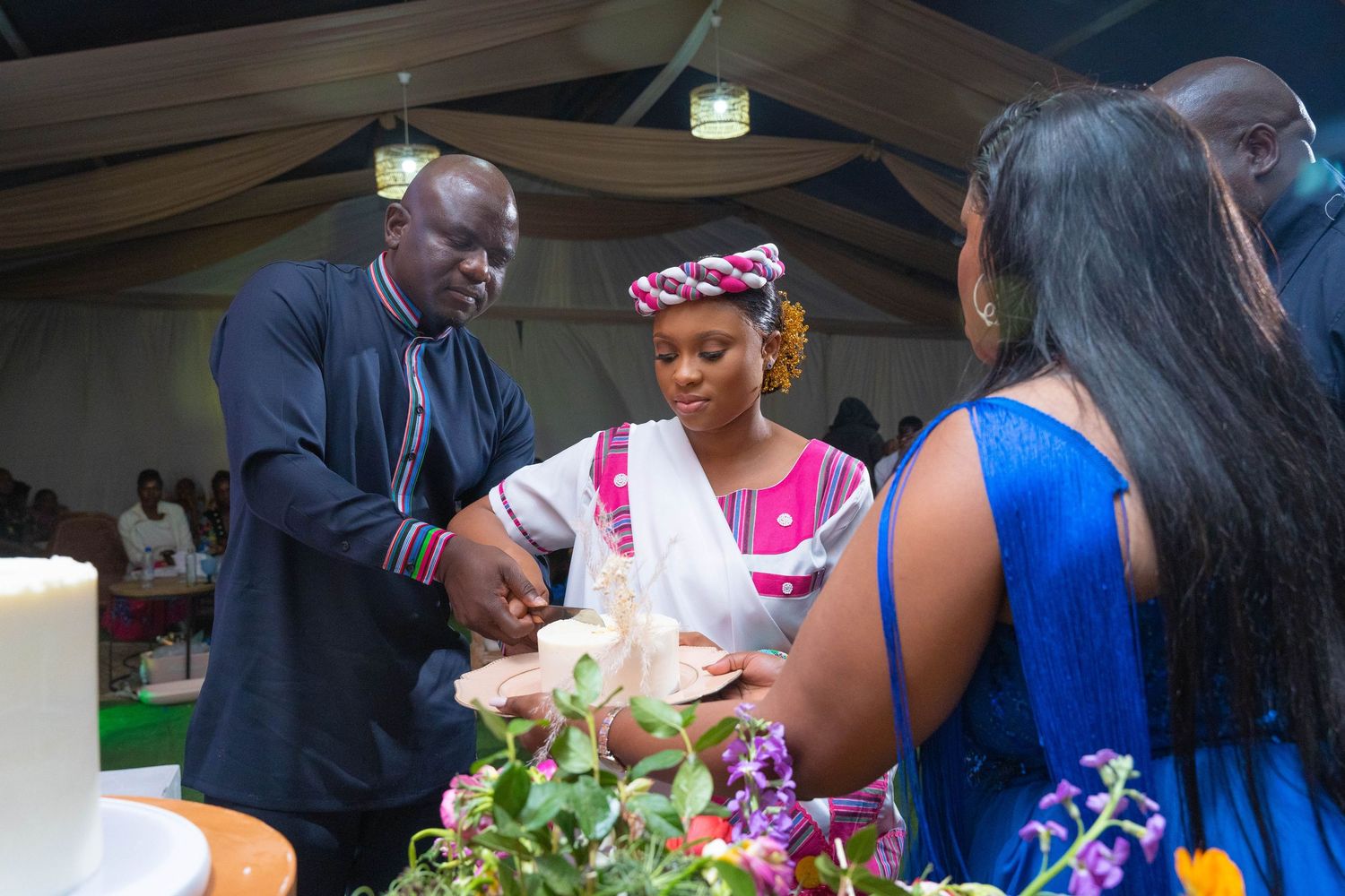 African couple cutting their wedding cake together assisted by a guest at a traditional wedding reception.