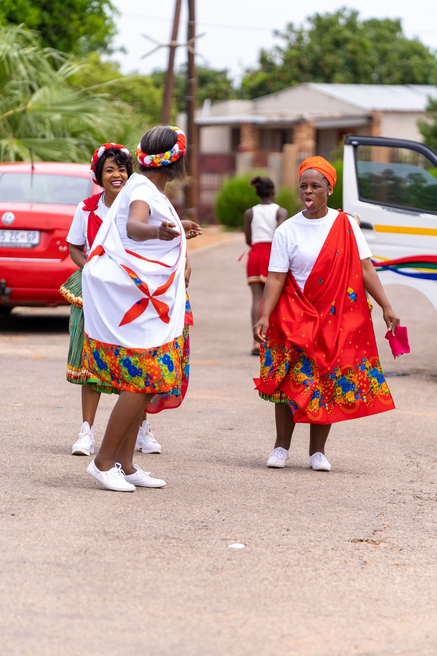 Three women in colorful traditional South African Xhosa attire dance together on a street during cultural celebration.