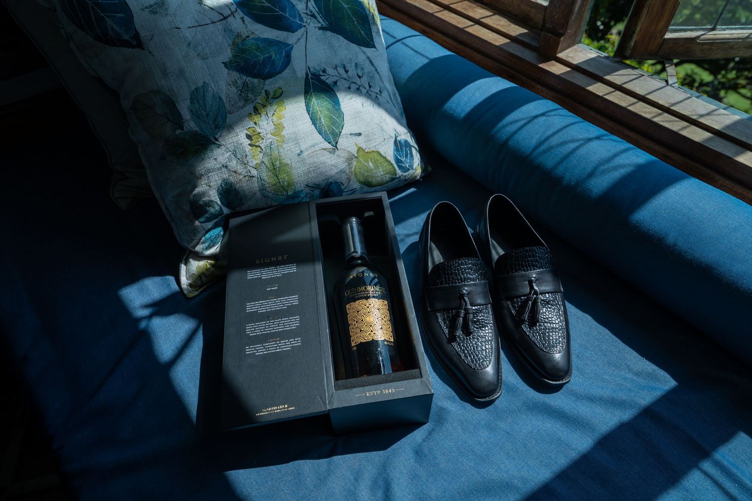 Black loafer shoes and a boxed bottle of wine on a blue sofa, wedding day groom accessories.