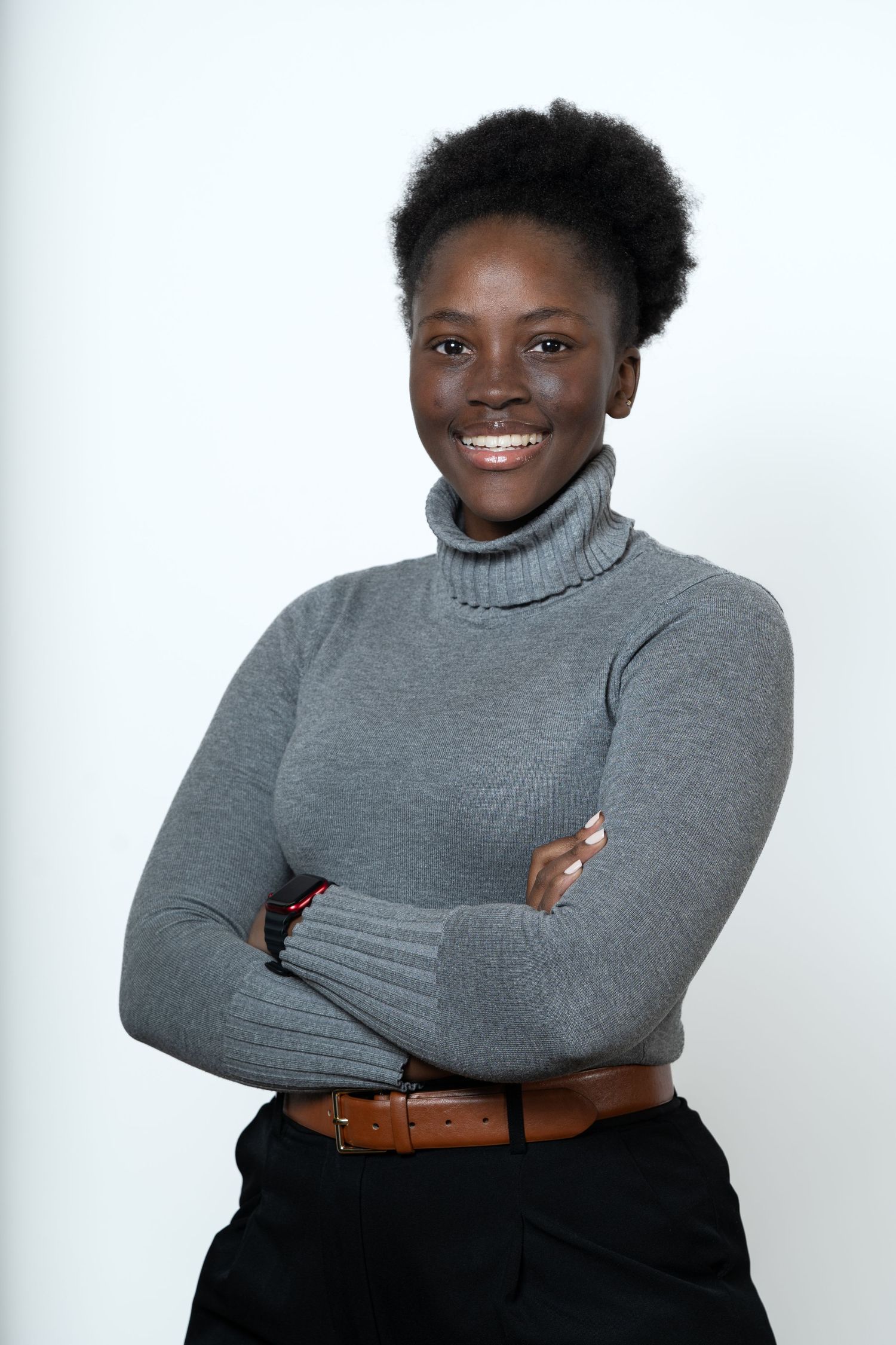 Smiling young Black woman in gray turtleneck sweater with arms crossed, posing confidently against white background.