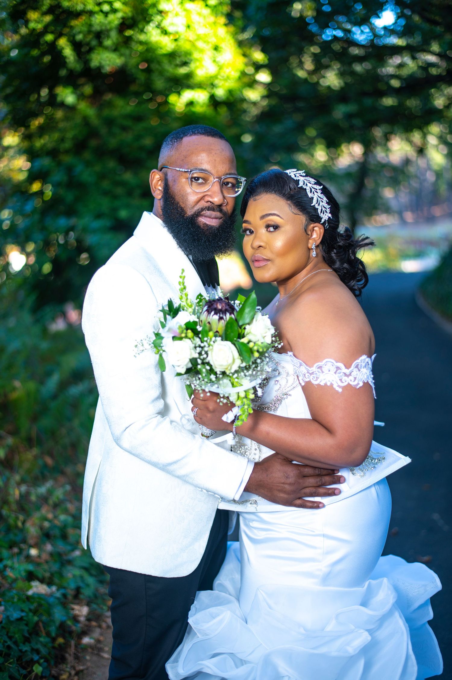 Bride and groom portrait, groom in white tuxedo holding bouquet, bride in lace off-shoulder mermaid gown in garden.