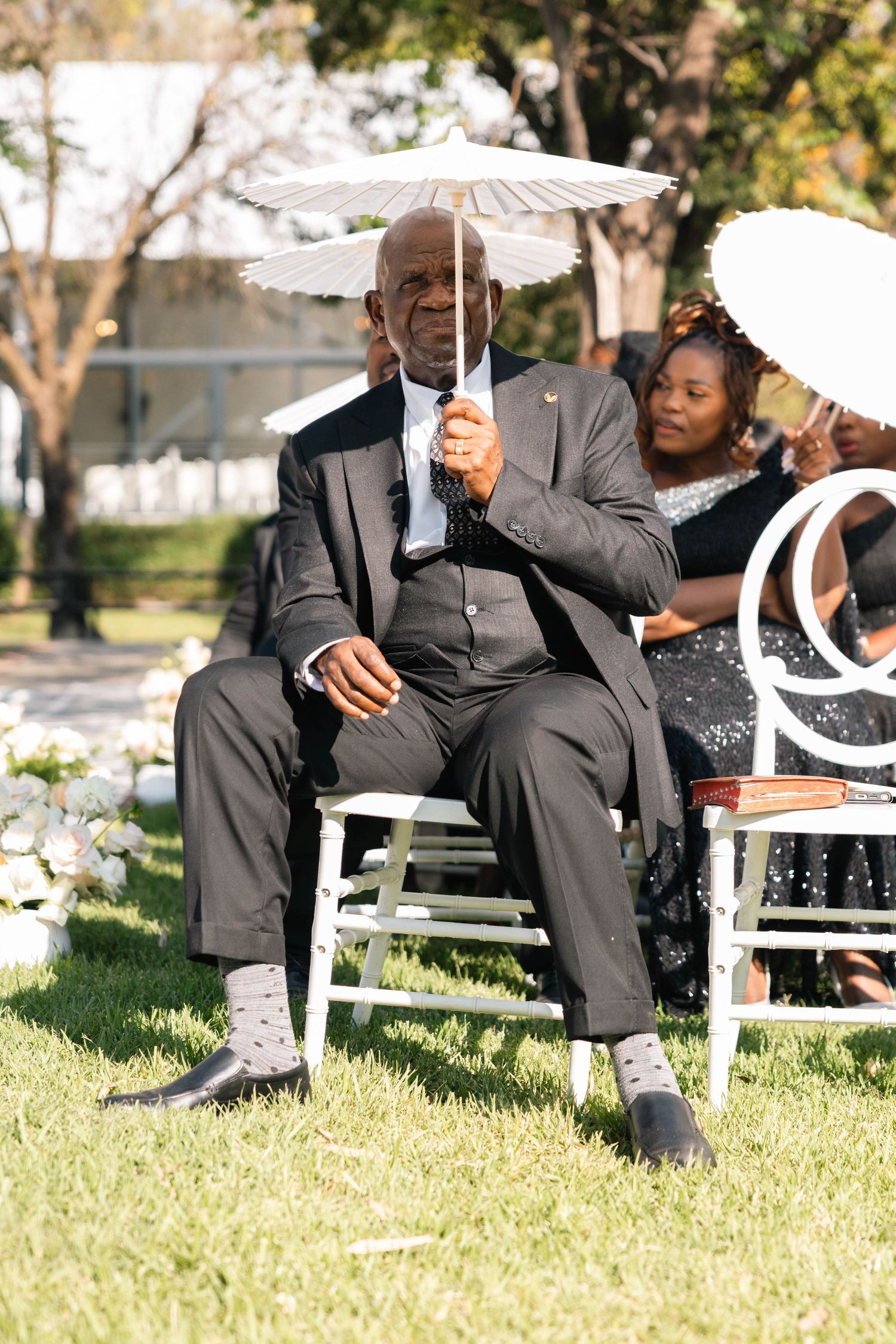 Elderly man in a dark suit holding a white parasol while seated at an outdoor wedding ceremony.