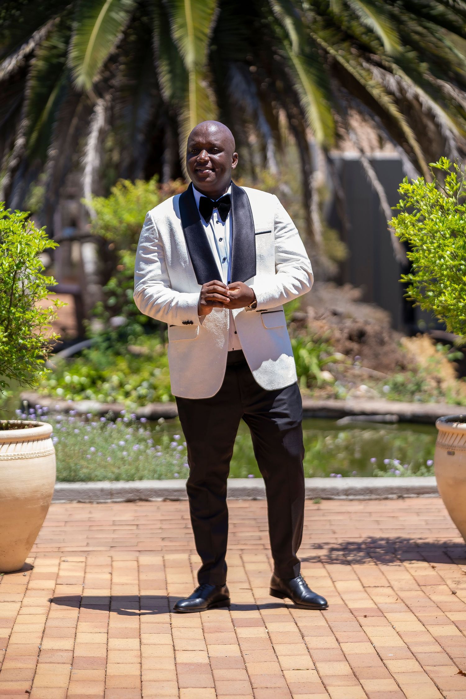 Groom in white tuxedo jacket and black bow tie posing in a lush garden setting.