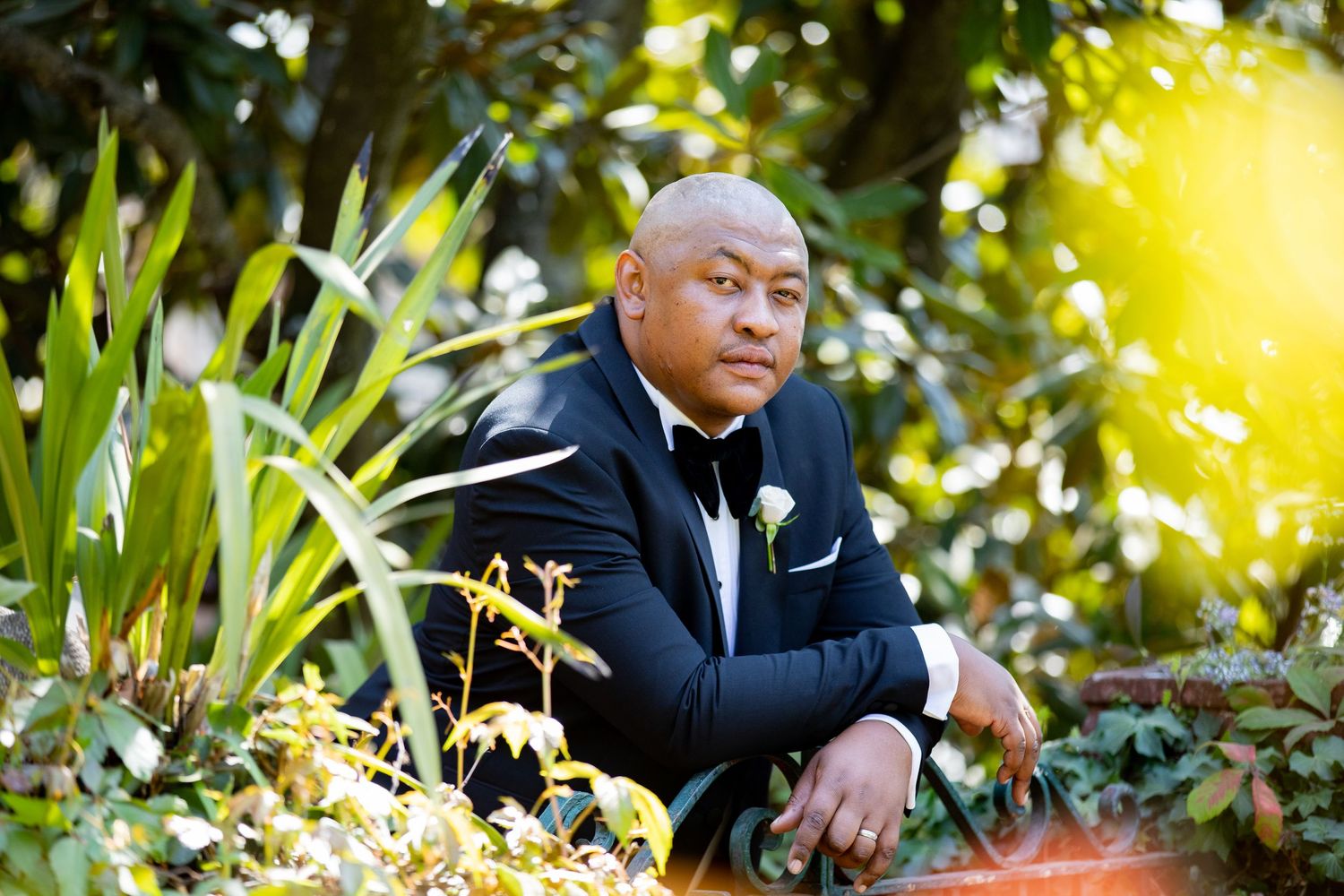 Groom in black tuxedo with white boutonniere posing outdoors among lush green garden foliage.